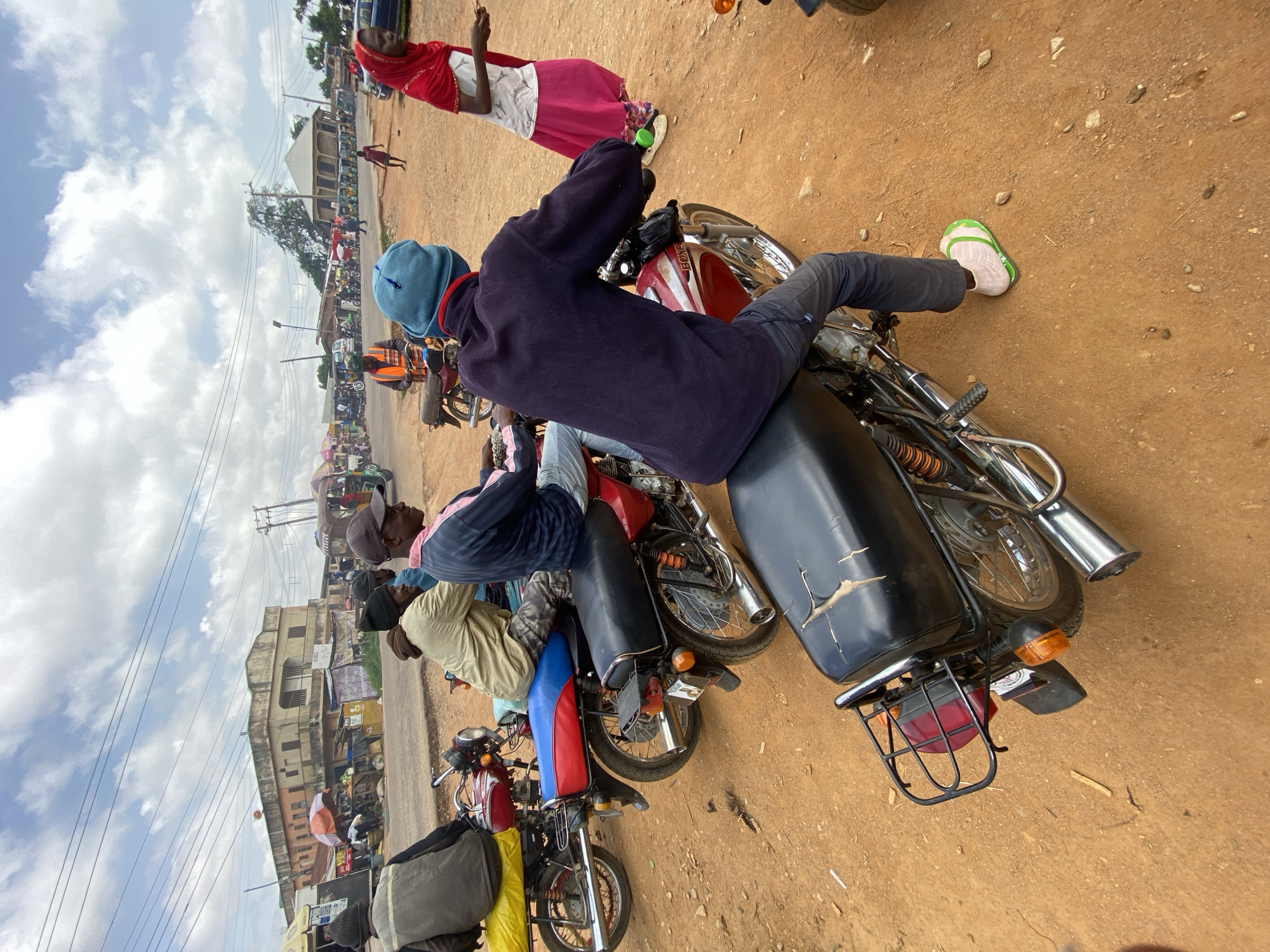 In Gwong, Nigeria, a dusty street scene unfolds under a cloudy sky. The foreground is dominated by a black motorcycle, its seat showing signs of wear, and the lower body of a person wearing dark pants and a light-colored sock with a green flip-flop. To the left and slightly behind, another motorcycle, red with a blue seat, is occupied by a person wearing a light-colored jacket. Further back, a third motorcycle, red with a yellow seat cover, has a person in a dark vest. In the mid-ground, a person in a brown jacket is on a motorcycle, and another individual in a blue jacket and cap is visible on a motorcycle. A woman in a red headscarf and a pink skirt stands to the right, holding something in her hand. In the background, a row of motorcycles and tricycles are parked along a street, with buildings and stalls lining the road. The ground is a mix of dirt and gravel.