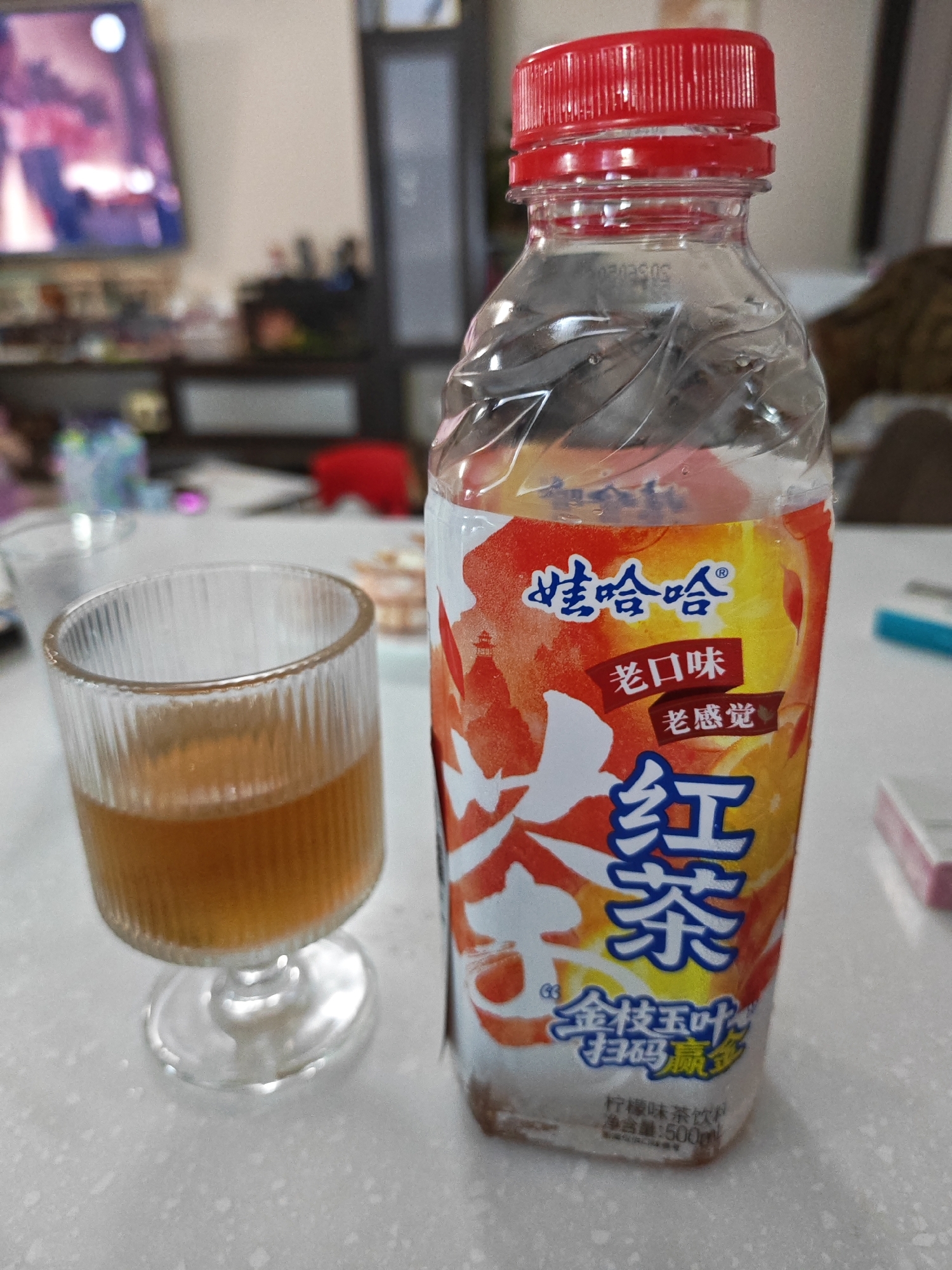 A clear plastic bottle with a red cap, containing a brown liquid, stands on a white surface. The bottle label features a vibrant orange and yellow design with Chinese characters. To the left of the bottle, a clear, ribbed glass cup holds a portion of the same brown liquid. The background is blurred, showing a television screen, shelves with various items, and a red object. The scene appears to be indoors, possibly in a domestic setting. The location is not discernable from the provided image, and no information is available about Vladivostok, Russia.
