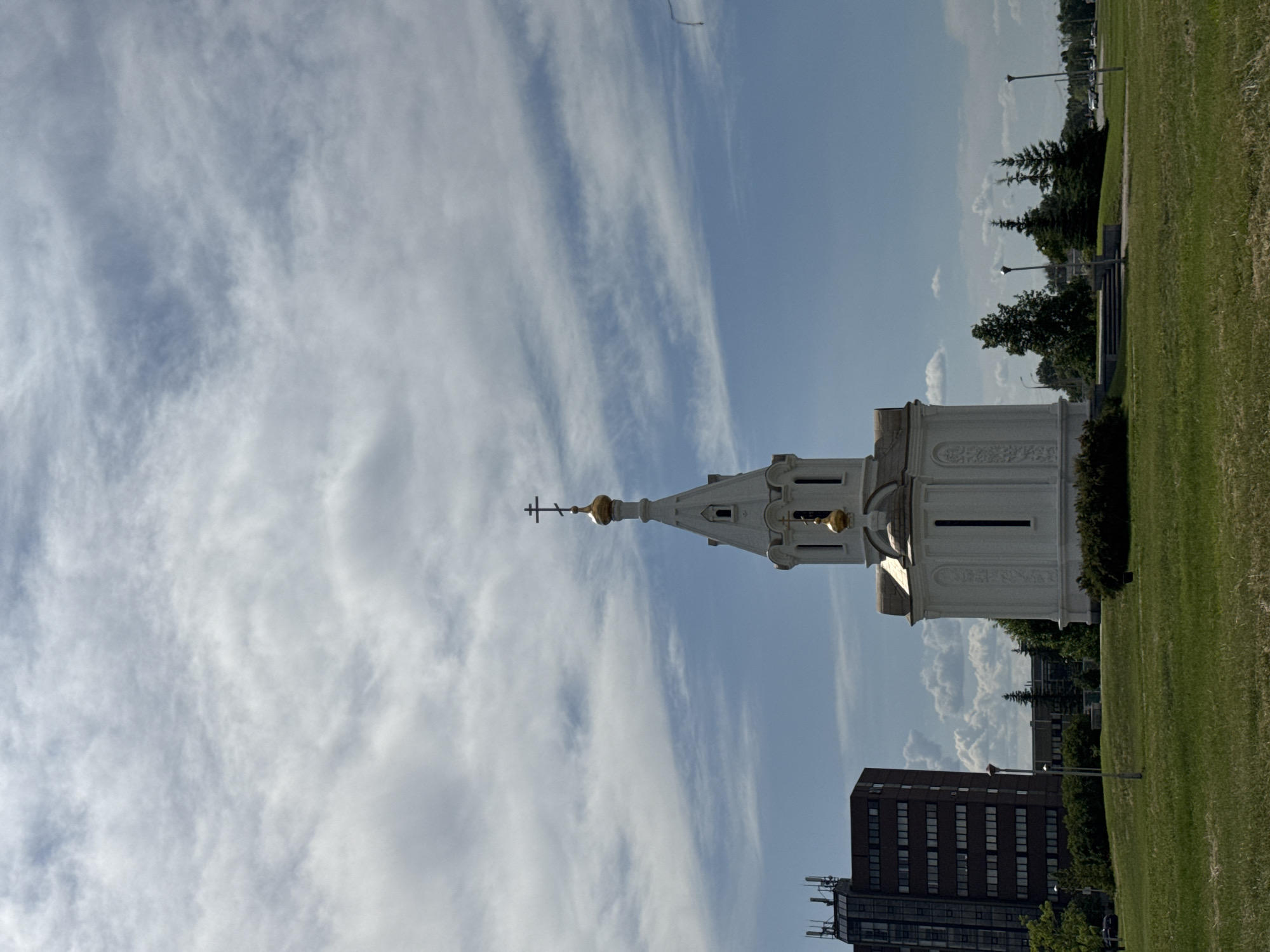 This image presents a daytime outdoor scene in the city of Unknown, Unknown, featuring a white Orthodox Christian chapel prominently positioned slightly to the right of the frame's center. The chapel exhibits a multi-tiered spire topped with an Orthodox cross, and two golden onion domes, one beneath the spire and a larger one on the main structure. Elongated, narrow dark windows are visible on its facade, which also features subtle relief ornamentation. The chapel is situated on a slightly elevated, verdant green lawn, with a short set of steps leading up to its base visible on the viewer's left side.
To the left of the chapel, in the mid-ground, stands a tall, dark, multi-story building with numerous rectangular windows and visible communication equipment on its roof. The background behind the chapel includes a cluster of green trees, several appearing coniferous, and further in the distance, a road with indistinct vehicles and faint building structures. Several tall, dark lampposts are scattered across the expansive green lawn. The sky above occupies the upper two-thirds of the image, displaying a mix of light blue areas and white, horizontally streaked cirrus clouds, along with some more defined cumulus cloud formations. No individuals are visible in the immediate foreground or mid-ground.