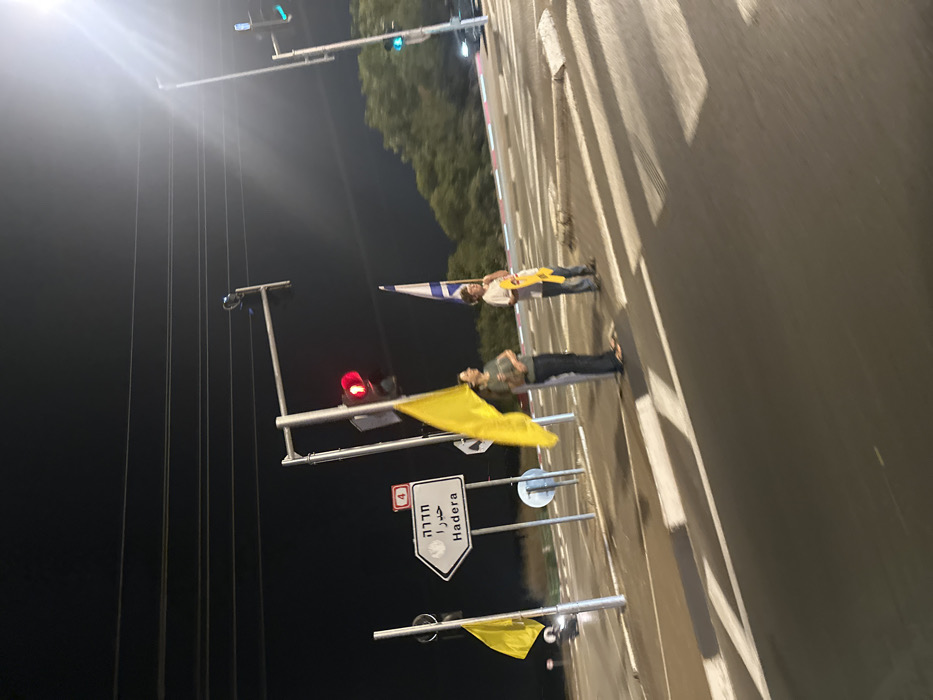 The image captures a nighttime scene at an intersection on Road 4 near Mishmar Hasharon, Israel, with a sign indicating the direction to Hadera. Two individuals stand on the paved shoulder next to a marked pedestrian crosswalk. The individual on the left, an adult, wears dark clothing and holds a large, bright yellow flag. To their right, a second individual, appearing younger, wears a light-colored shirt and jeans, holding an Israeli flag and a smaller yellow flag or sign. A third yellow flag is attached to a pole further to the left. Overhead, power lines span across the dark sky. Traffic infrastructure includes a white directional sign for "Hadera" and "4," multiple streetlights, and traffic signals; one signal displays a red light in the foreground, while another in the distance shows green. The roadway features white pedestrian crossing stripes and red-and-white painted curbstones. Vehicle lights are faintly visible in the far background on both sides of the road.