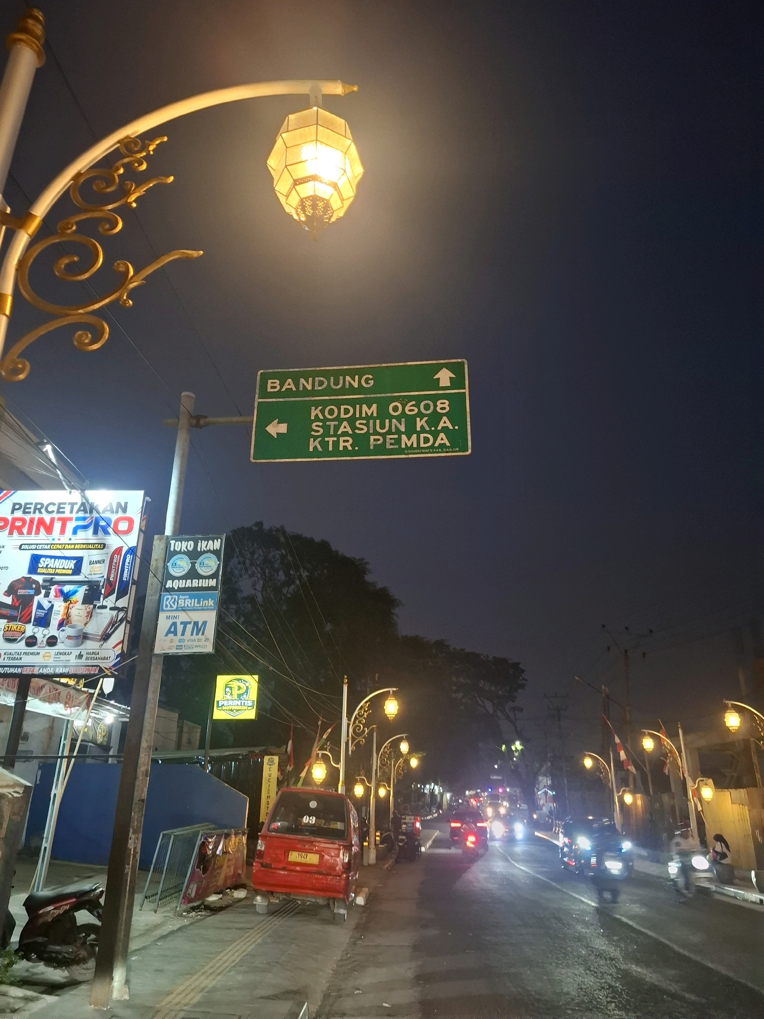 The image depicts a street scene at night in the city of Sayang, Indonesia, illuminated by decorative streetlights and various commercial signs. A green directional sign with white text indicates "BANDUNG" with an upward arrow, and "KODIM 0608 STASION K.A. KTR. PEMDA" with a leftward arrow. To the left, a large advertisement for "PRINTPRO" is prominently displayed, showcasing various printing services and products. Below it, a smaller sign for "TOKO IKAN AQUARIUM" is visible, along with an ATM sign for "BRI Link". A red three-wheeled vehicle is parked on the side of the road, facing away from the camera. Several motorcycles are also present, one parked on the sidewalk and others on the street. The street is wet, suggesting recent rain, and vehicles with their headlights on are driving down the road. The environment appears to be a commercial or urban area, with buildings and trees visible in the background. The overall atmosphere is that of a bustling nighttime street in Indonesia.