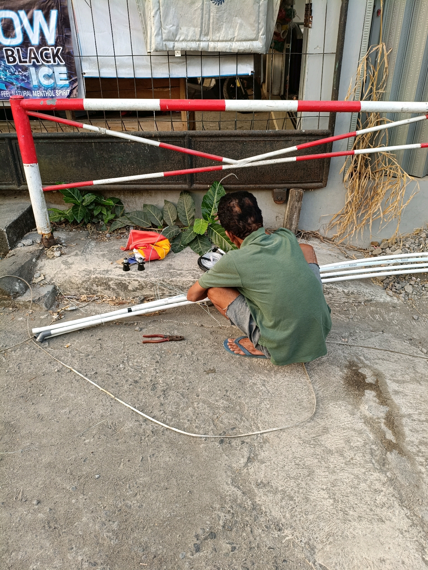 A person is crouched on a concrete surface in Maguwoharjo, Indonesia, working with white PVC pipes. The person, wearing a green t-shirt and grey shorts, is surrounded by various materials including a pair of red pliers, what appears to be a coil of wire, and a colorful fabric bundle. In the background, a red and white striped barrier is visible, possibly a gate or a construction barrier, along with some foliage and a dark-colored corrugated metal surface. The environment suggests an outdoor urban setting, with the concrete ground showing signs of wear and tear, and scattered debris. The overall scene depicts a task being carried out in a seemingly public or semi-public outdoor space.