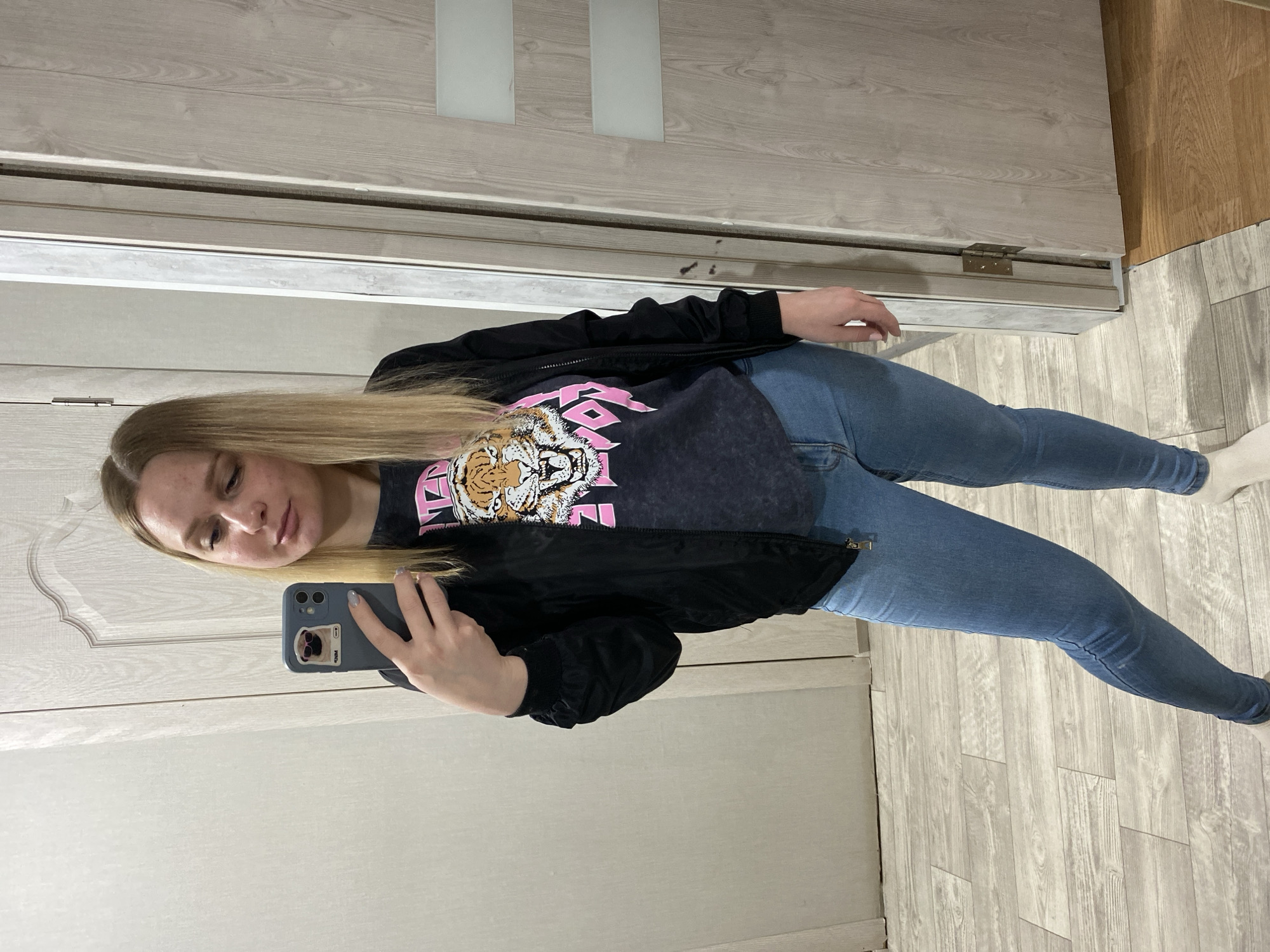 A young blonde woman with fair skin takes a mirror selfie in a room with light-colored wooden floors and doors. She is wearing a dark gray t-shirt with a large graphic of a roaring tiger's head in pink and orange, a black bomber jacket, and light blue skinny jeans. She is holding a dark gray smartphone in her left hand, which displays a sticker of a pug wearing sunglasses on its case. Her hair is shoulder-length and straight. The room appears to be a hallway or a dressing area, with a closet door and another door visible in the background.