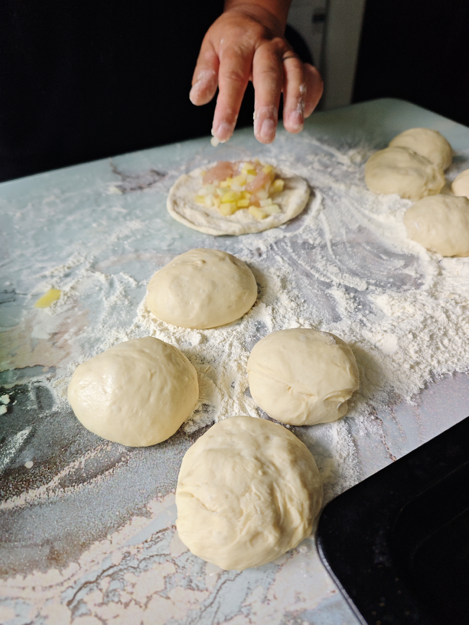 A hand hovers over a dough circle filled with diced ingredients, on a light blue marbled countertop dusted with flour, surrounded by several dough balls. A black object is visible at the bottom right corner. The image does not provide sufficient information to confirm a specific location in Фоки, Russia.