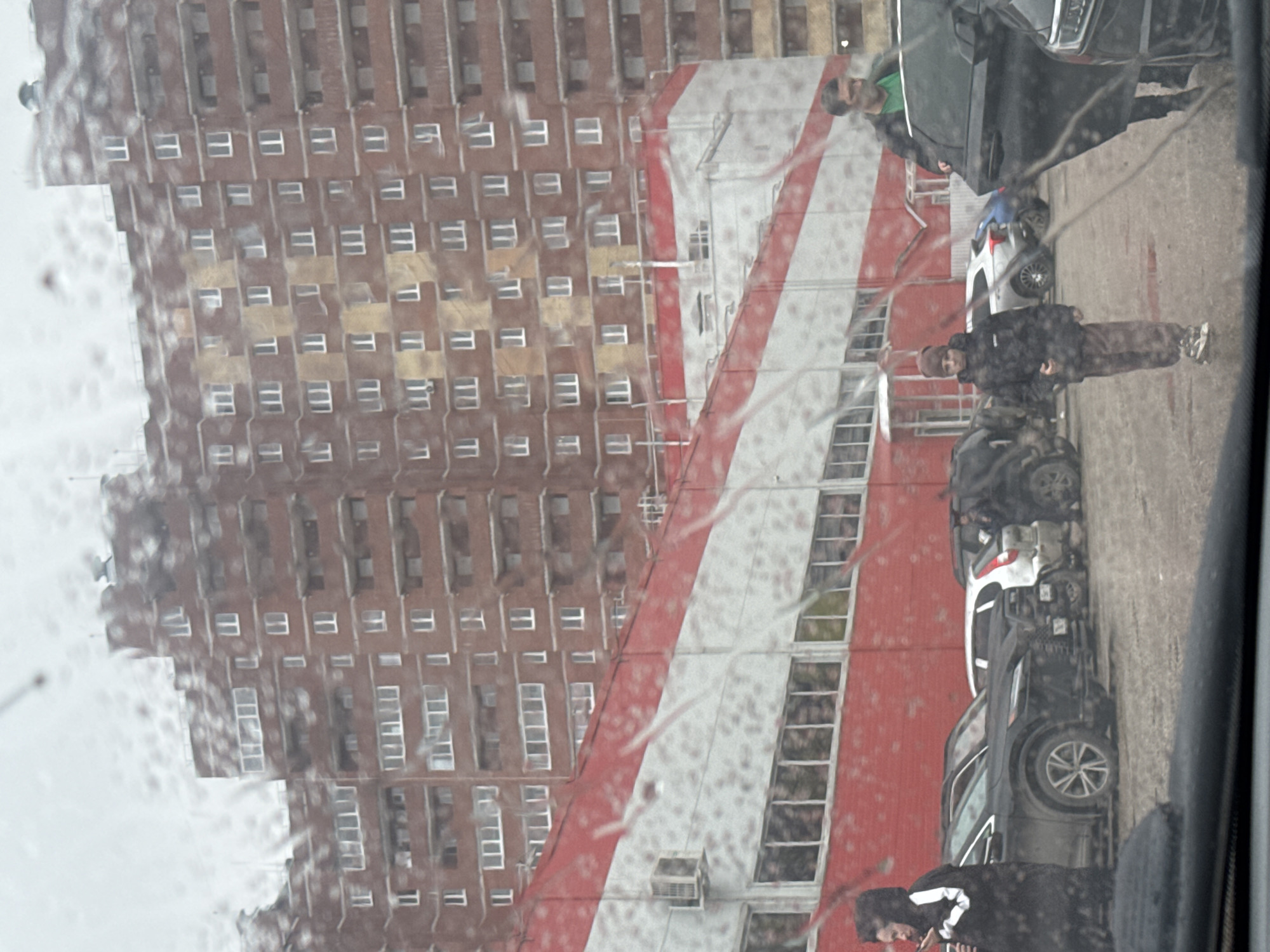 The image, taken through a rain-streaked window, depicts an urban scene in Perm, Russia, under overcast conditions. A tall, multi-story brick building with numerous windows occupies the background. In front of it, a smaller building features a distinctive facade with diagonal red and white stripes, and several air conditioning units are visible on its roofline.
A paved area in the middle ground serves as a parking lot or street, with several cars parked. A dark SUV is prominent in the foreground. Four individuals are visible. On the left, a person wearing a black hooded jacket with white markings appears to be looking down. Further back, another individual in a dark jacket and brown pants stands near the black SUV, looking towards the camera. A third person, wearing a green shirt and dark jacket, is partially visible behind the SUV, also looking towards the viewer. A fourth person is partly seen near a white car in the background.
The water on the camera lens creates a blurred and streaky effect across the entire image, obscuring fine details and suggesting rainy or damp weather. The time of day appears to be daytime, though the light is diffused due to cloud cover. No specific activity or emotion is clearly discernible, and no visible text is legible.