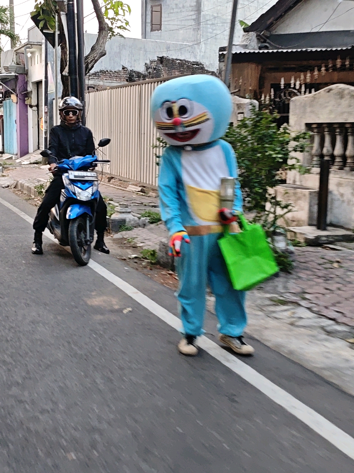 A person in a Doraemon costume walks on a street in Kediri City, Indonesia. The Doraemon character is blue and white with yellow accents, and is holding a green shopping bag and a cylindrical object in its hands. The person is wearing light brown shoes. Behind the Doraemon character, a person in dark clothing and a helmet rides a blue scooter on the same street. The street is paved and has a white lane marker. The surrounding environment includes buildings with light-colored walls and some greenery. The image is taken from a slightly elevated perspective, looking down at the street.