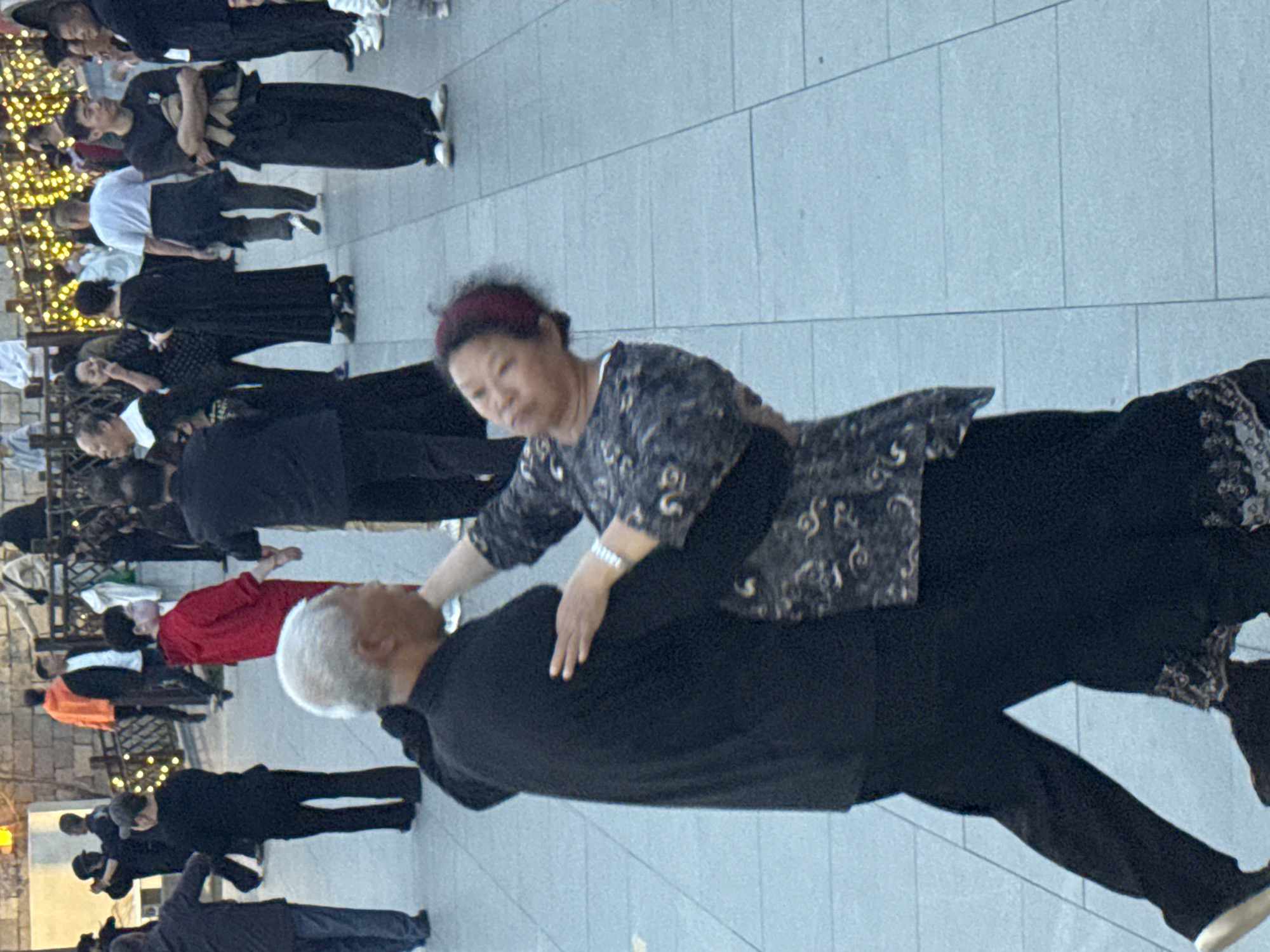 A medium shot depicts an outdoor scene in Jinzhou, China, with a group of people engaged in a synchronized activity. In the foreground, an older Asian woman with short, dark hair and a red headband is positioned behind a silver-haired Asian man wearing a black shirt and pants. She has her hand on his back. They are surrounded by other individuals, most of whom are dressed in dark-colored clothing.
The group is arranged in rows, with some individuals in red shirts and others in white tops. The background includes urban elements, such as paving stones. A string of lights is visible in the upper left corner, suggesting a possible festive or decorative context.