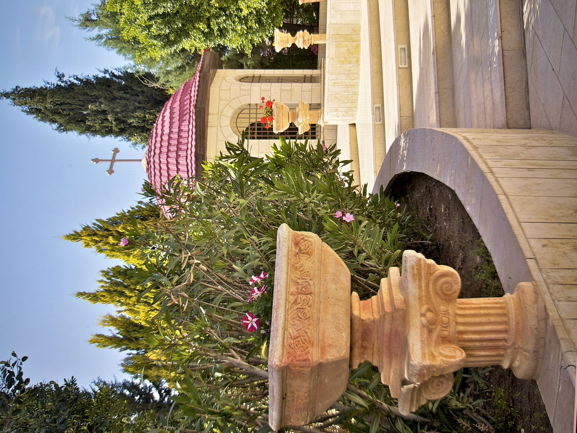 The image depicts an outdoor scene featuring a small, ornate chapel with a pink, tiled, domed roof topped by a cross. The chapel is constructed of light-colored stone and has arched windows with decorative metal grilles. In front of the chapel, there are stone steps and planters. A large, elaborate stone urn, possibly a fountain base or planter, with carved decorative motifs sits prominently in the foreground to the left. Behind the urn, lush green foliage and trees are visible, including tall, slender cypress trees. The ground is paved with stone tiles, and a curved stone wall lines the foreground. The sky is a clear blue. This scene is located at the St. Sophia's Cathedral complex in Kyiv, Ukraine, specifically in the area of the refectory church. There are no people or animals visible in the image.