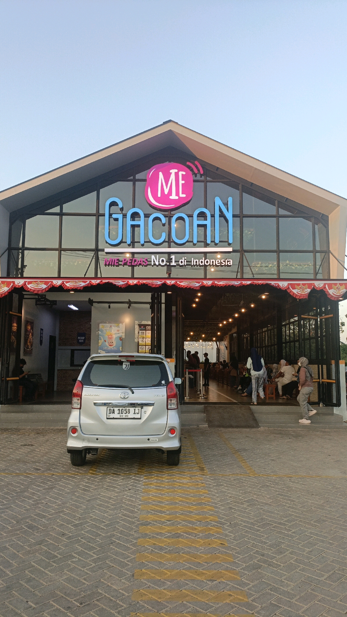 The image shows the exterior of a restaurant called "MIE GACOAN" located in Pelaihari, Indonesia. The restaurant features a modern architectural design with a large glass facade and a prominent sign with the restaurant's name. The sign is illuminated with blue neon lights and a pink circular logo with the letters "ME". Below the main sign, smaller text reads "MIE PEDAS No. 1 di Indonesia".
In front of the restaurant, a silver minivan is parked in a designated parking spot marked with yellow lines on a brick-paved area. Several people are visible entering and exiting the restaurant, some are seated at tables inside, and others are standing near the entrance. The interior of the restaurant appears to be well-lit with decorative lights, and there are patrons dining at tables. The overall atmosphere suggests a busy and inviting dining establishment. The sky is clear and blue, indicating it is daytime.