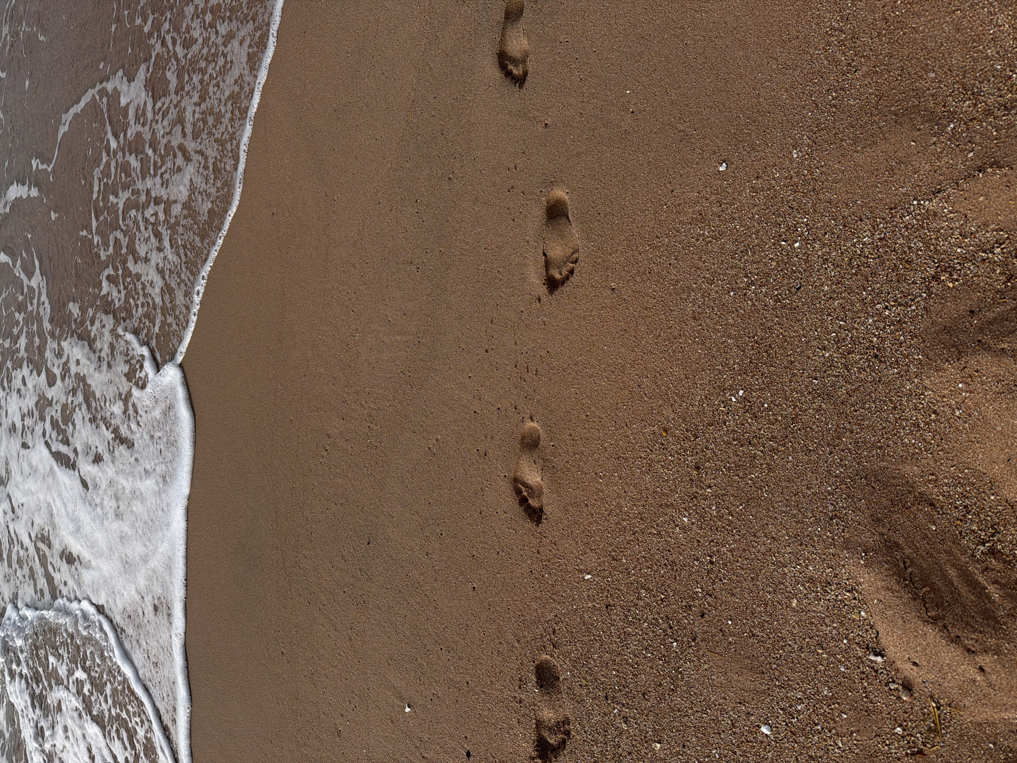The image depicts a beach scene. White foamy waves are on the left side of the image meeting the sand. A line of footprints extends away from the water along the sand. The sand gradually changes texture, from fine to more pebbly and scattered with small shells. The location is likely a beach in Sharm El Sheikh, Egypt, judging from the sand color and wave characteristics. No people are visible. The action depicted is the tide encroaching on the shore and the presence of footprints indicating past human activity.