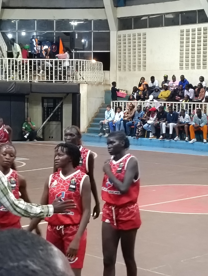 A group of female basketball players wearing red and white jerseys with a diamond pattern stand on the court. Spectators are visible in the background, seated on bleachers and standing on a balcony. The game appears to be indoors, with lighting illuminating the court and surrounding areas. Some individuals in the crowd are wearing casual clothing, while others are dressed more formally.