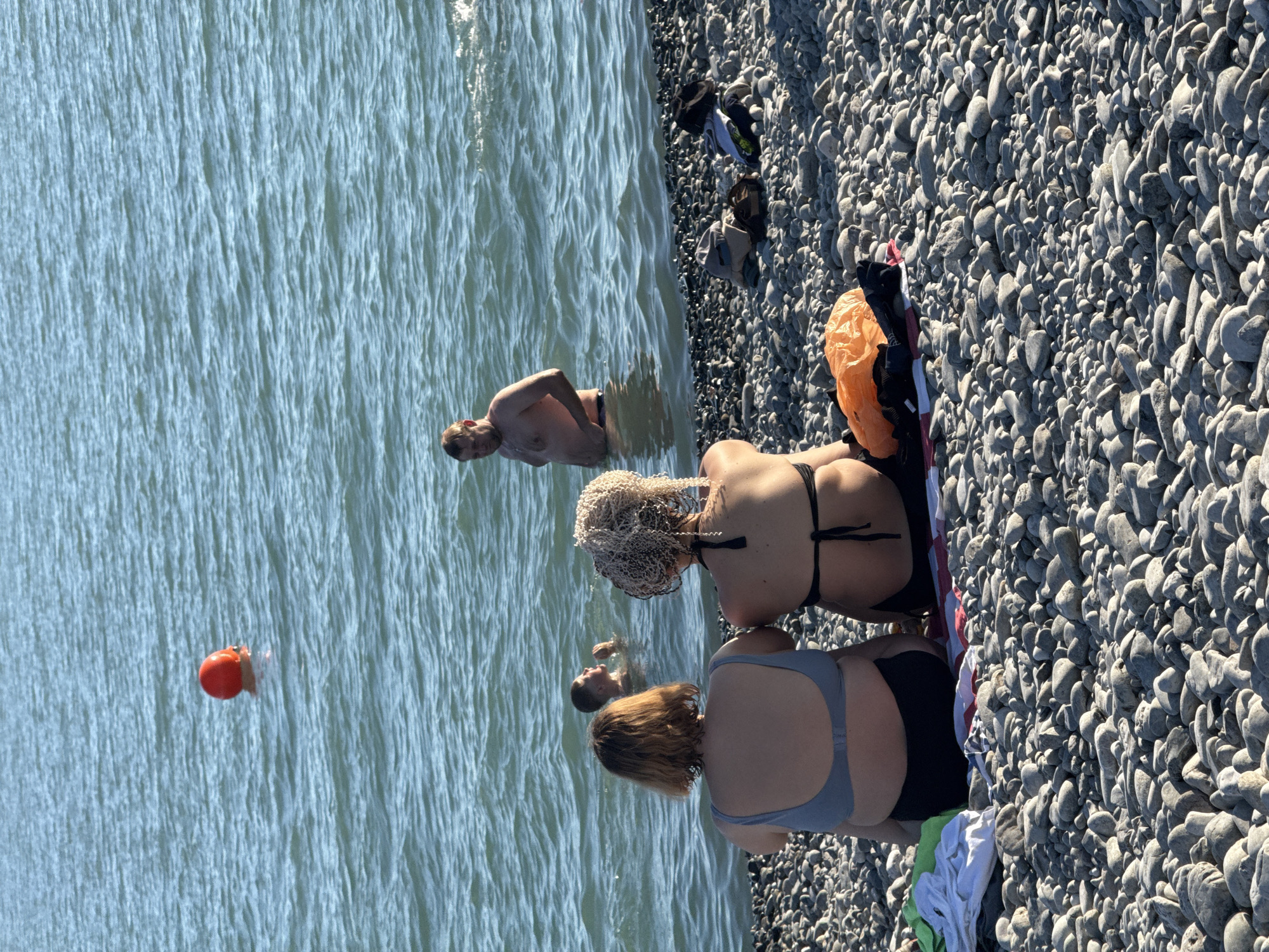 The image displays a daytime scene on a pebble beach in the city of Sirius, Russia. The foreground features two women seated on the grey, rounded pebbles with their backs to the viewer, facing the sea. The woman on the left wears a light grey or blue bikini top and dark bottoms, sitting on a green and white fabric. The woman on the right wears a black string bikini top and dark bottoms, with voluminous blonde braided hair; she is seated on a red and white striped fabric. An orange plastic bag and a dark bag are situated on the pebbles near the right woman.
In the mid-ground, three men are partially submerged in the blue-green, rippling water. To the right, a man stands chest-deep, shirtless, facing slightly left. To the left, another man is partially submerged, appearing to look upwards or to the side. Further out to the left, a person's head, largely obscured by a bright orange swimming cap, is visible above the waterline. On the pebble beach to the far right, several items of clothing and shoes are casually placed. The overall lighting suggests bright sunlight.