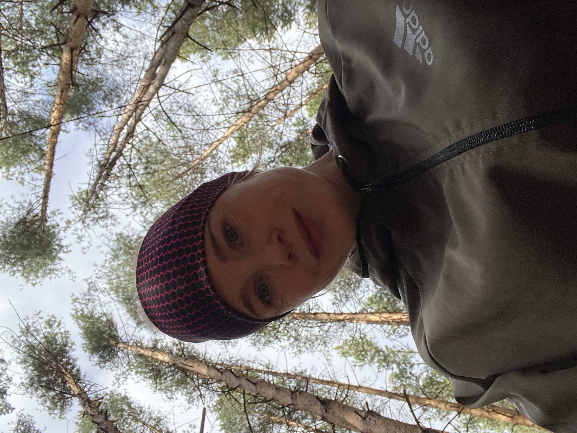 A woman is taking a selfie from a low angle while standing in a forest. She is wearing a dark-colored Adidas jacket and a dark headband with a pink honeycomb pattern. Pine trees surround her, with branches extending upwards towards the sky. The sky is visible through the trees. The location is implied to be in the city of Chaikovsky, Russia.