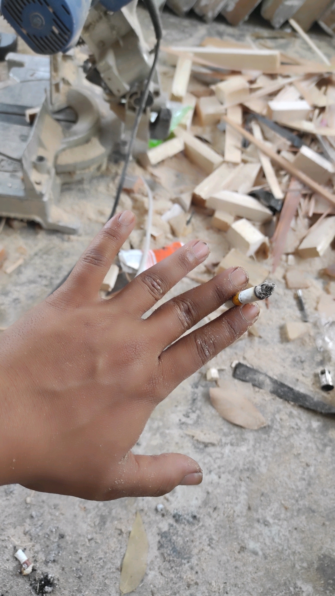 A close-up, over-the-shoulder shot captures a hand holding a lit cigarette amidst a construction site. The hand, lightly dusted with sawdust, is positioned in the foreground. The cigarette, held between the index and middle fingers, shows a glowing ember. In the background, a chop saw is visible, along with scattered wood scraps, tools, and debris, suggesting an active work area. The ground appears to be concrete or a similar rough surface, also covered in sawdust and small pieces of wood. The overall impression is one of a brief pause in manual labor. The scene is likely located in Guwosari, Indonesia, given the context.
