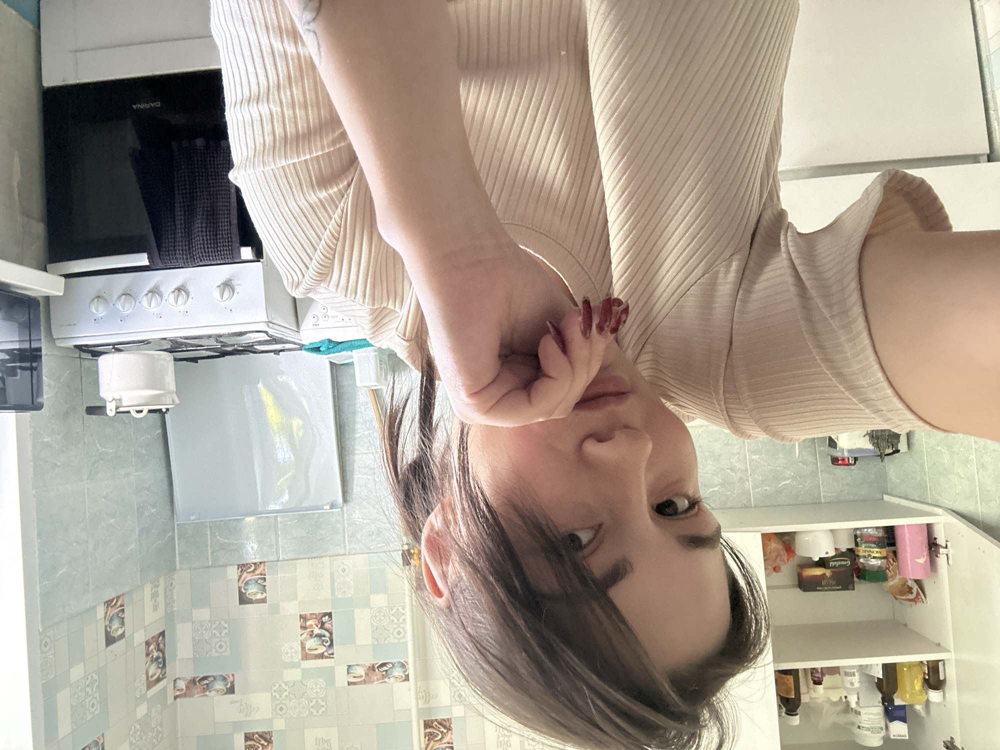 A young woman with dark hair is taking a selfie in a kitchen. She is wearing a beige, ribbed t-shirt and has her left hand resting on her chin. In the background, a white stove with a black oven door is visible, along with kitchen cabinets and patterned wallpaper. The woman appears to be in an indoor environment, likely a residential kitchen.