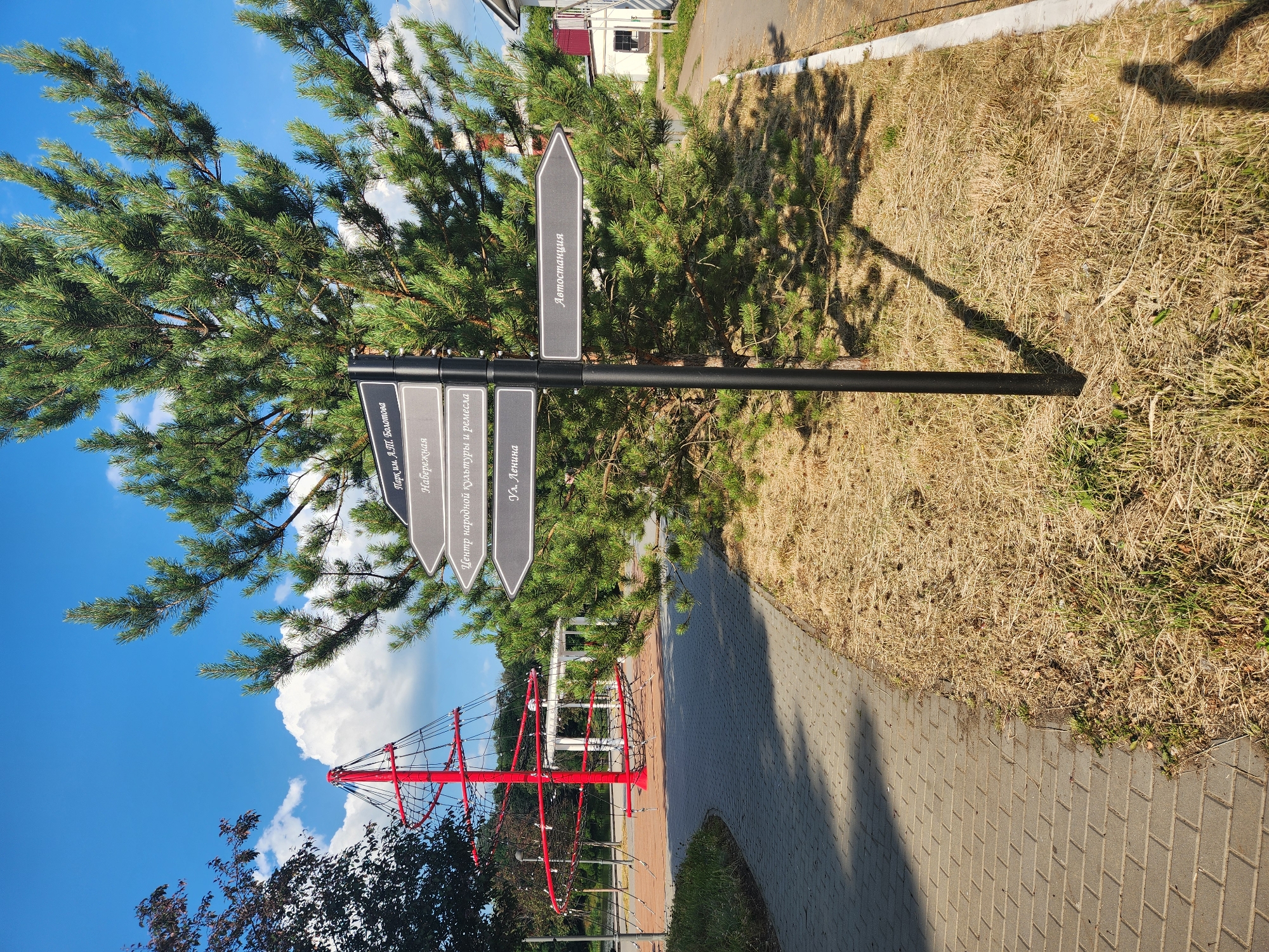 This image shows a signpost with directional arrows in a park-like setting. The signpost is black and has multiple grey arrows pointing in different directions. Each arrow has text written on it in a lighter font. The visible text on the arrows includes "Парк им. А.Т. Болотова", "Набережная", "Центр народной культуры и ремесла", "Ул. Ленина", and "Автостанция".
The signpost is positioned in front of a large pine tree with green needles. Behind the tree, there is dry, yellow grass and a paved walkway. In the background, a building with a red roof and a white section can be seen, along with a blue sky with fluffy white clouds. To the left of the signpost, a tall red structure with netting, possibly part of a playground or a decorative element, is visible against the blue sky and some dark green trees.
The lighting suggests it is daytime, likely sunny, given the shadows cast by the signpost and the tree. The dry grass indicates a warm season, possibly summer or early autumn. The overall scene depicts an outdoor public space, likely a park or recreational area, with signage providing directions to various points of interest within or near the park. There are no people visible in the image.