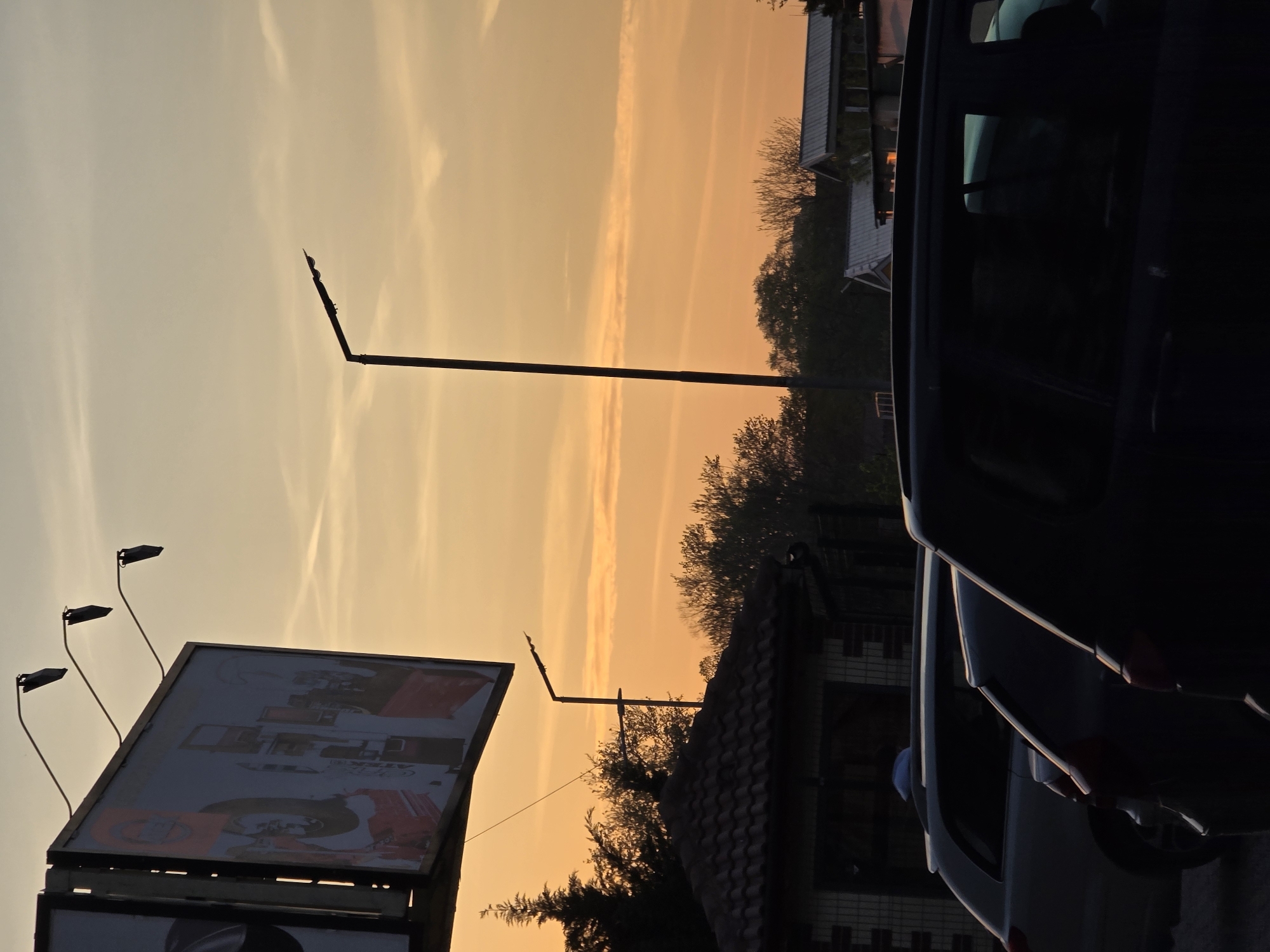 The image captures an evening scene in Plovdiv, Bulgaria, marked by a warm, orange-hued sunset. A large billboard with a visible advertisement is on the left. Behind the billboard, three curved lampposts extend upwards. A taller, straight lamppost rises near the center of the frame, extending beyond the rooftops of nearby buildings, including one with a tiled roof. Deciduous trees add texture to the skyline. The right side of the image is dominated by the dark silhouette of parked cars, with reflections of the sunset visible on the car windows.