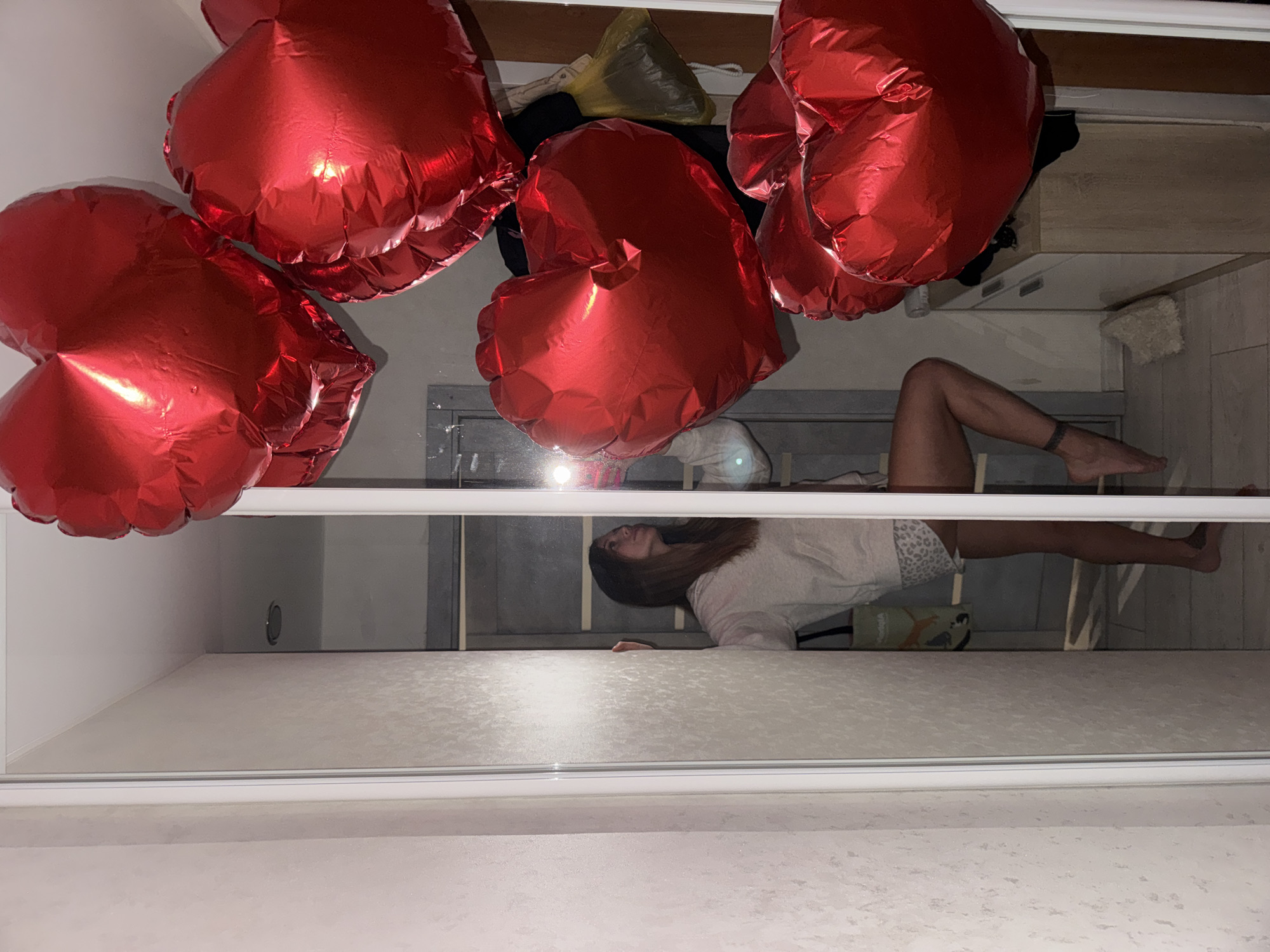 The image depicts a reflection in a mirror. Red heart-shaped balloons are clustered near the top left of the frame. In the mirror's reflection, a woman is lying down in what appears to be a room. The room's features are visible in the reflection, including a gray wall, flooring, and furniture. The image does not provide enough information to confirm the location or any specific actions/interactions taking place. The location in the city of Chaikovsky, Russia, cannot be confirmed.