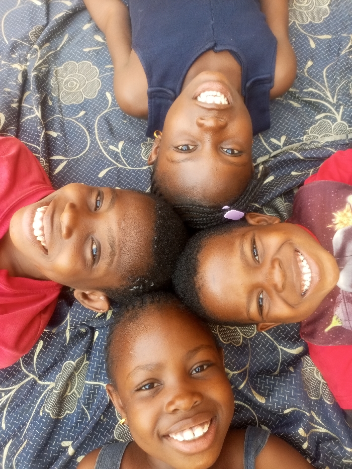 Four young girls are seen smiling brightly in a high-angle shot, arranged in a circular formation. They are lying on their backs with their heads meeting in the center of the frame, looking up directly at the camera. All the children appear to be of African descent, with dark complexions and dark, braided or short hair. Each girl displays a wide, joyful smile, revealing white teeth, conveying a strong sense of happiness and playfulness.
The child at the top of the frame wears a dark blue sleeveless top and small gold-colored hoop earrings. To the left, a child wears a red t-shirt, also with a bright smile. On the right, another child in a red t-shirt (with a partial graphic visible on the shoulder) has a purple hair tie and a radiant smile. The child at the bottom wears a dark grey or black sleeveless top and small earrings.
They are positioned on a patterned surface, likely a bed or mat, which features a dark blue or black background adorned with light-colored floral and vine-like designs. The scene is illuminated by natural, bright lighting, indicating it is daytime. The overall mood is cheerful and heartwarming. This image captures a happy moment in Tempure, Nigeria. There is no visible text in the media file.