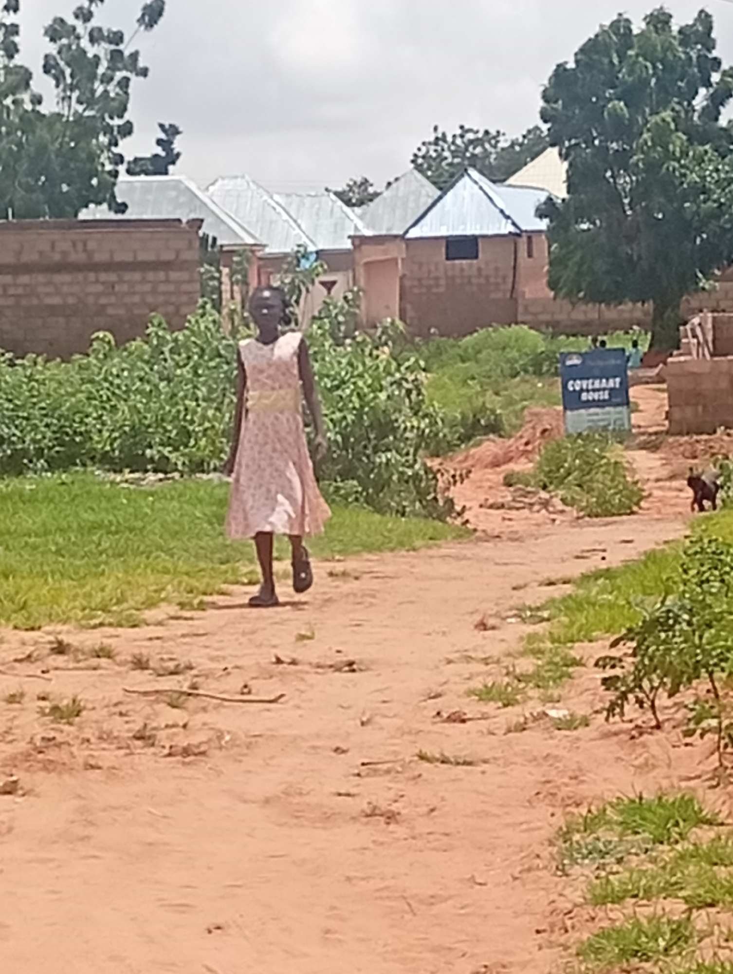 In Tumfure, Nigeria, a young girl with dark skin and dark hair is walking along a dirt path. She is wearing a light-colored dress with a floral pattern and a yellow sash. Her left leg is forward, and her arms are slightly bent at the elbows. Behind her, on the right side of the path, a small dark-colored animal, possibly a dog or a cat, is visible. The path is flanked by green grass and vegetation. In the background, there are buildings with brick walls and corrugated metal roofs, and several trees. A blue sign with white lettering that reads "COVENANT HOUSE" is also visible. The sky is overcast.