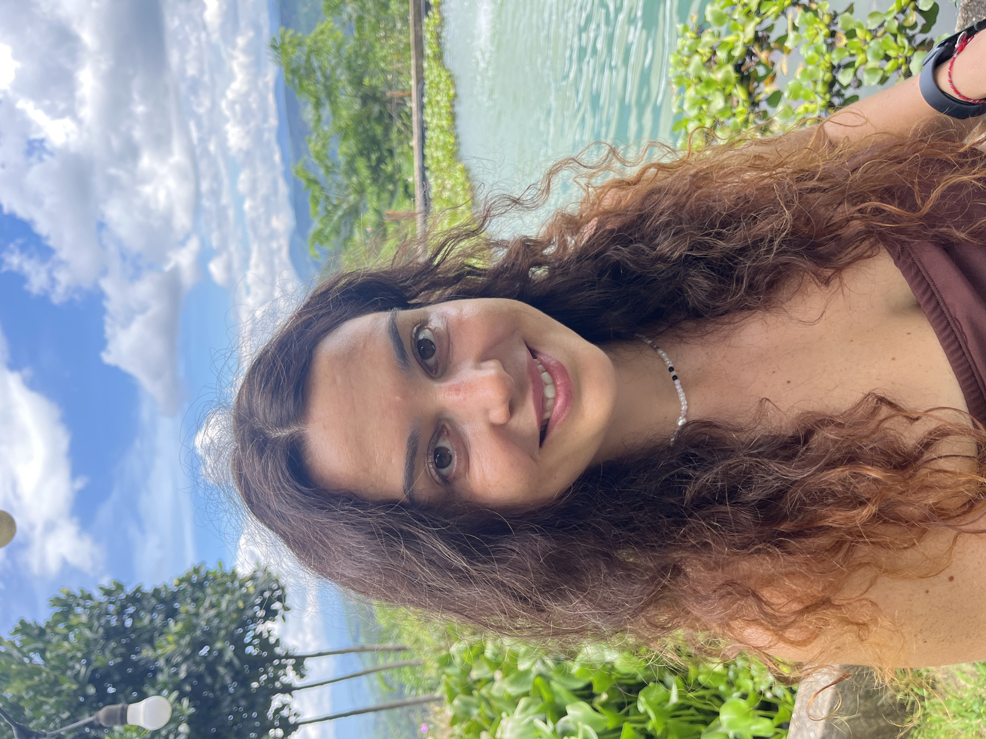 A medium shot captures a young woman with long, curly brown hair, smiling at the camera. She is wearing a brown top and a beaded necklace. The background suggests an outdoor setting, possibly a resort or natural park. Lush green vegetation and a body of turquoise water with lily pads are visible. In the distance, rolling hills are covered in trees, under a partly cloudy blue sky. The overall scene evokes a sense of relaxation and natural beauty, with a hint of an exotic location.