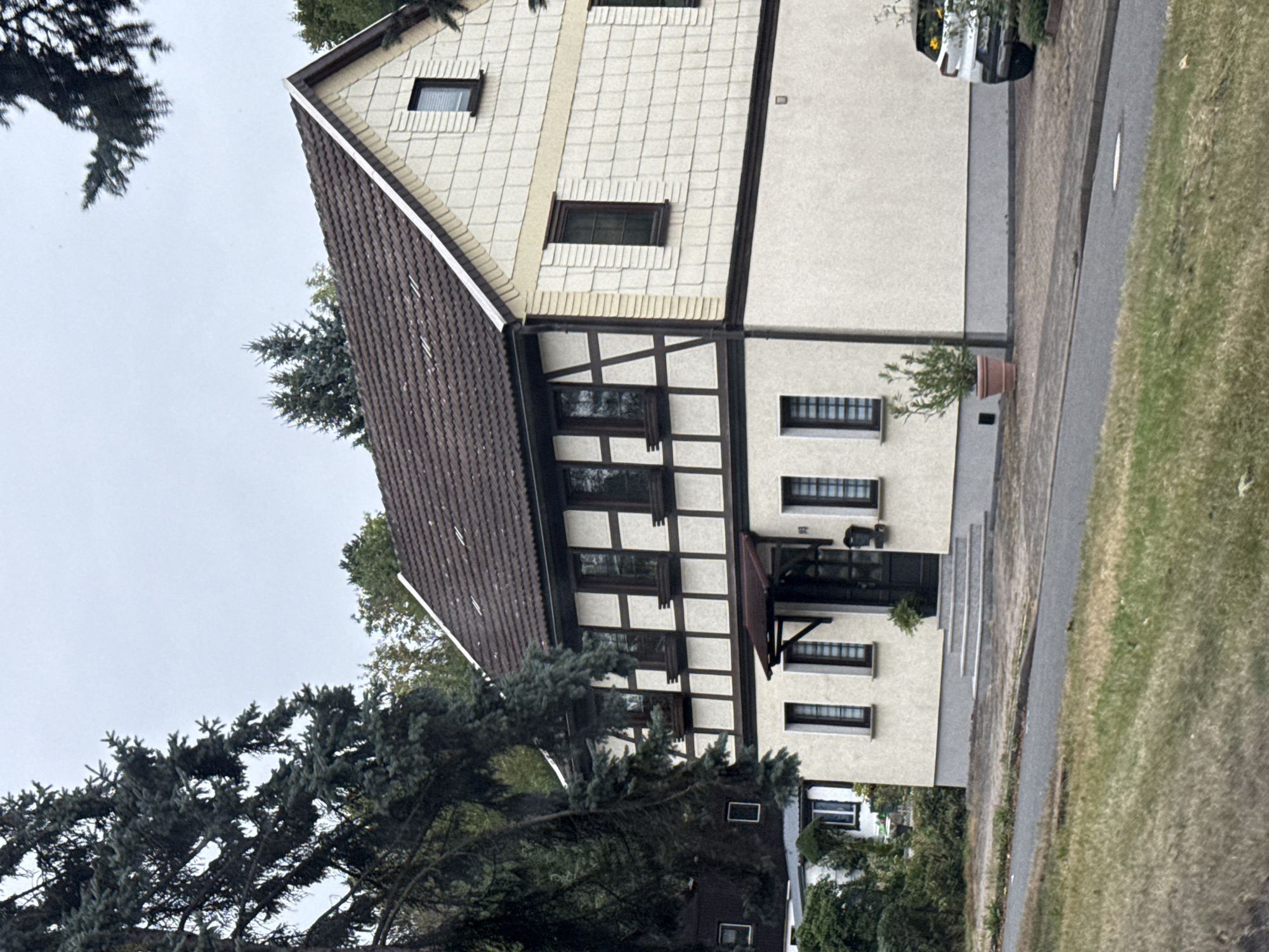 The image shows a two-story house with a brown tiled roof and a light-colored facade. The upper story features half-timbered detailing with dark brown beams and light-colored infill panels. Multiple windows are visible on both stories. A small, dark brown awning extends over the entrance. There are steps leading up to the front door. A potted plant sits to the right of the entrance. In front of the house, a paved driveway leads to a white SUV parked on the right side of the image. To the left of the house, another building is partially visible. The area in front of the house and driveway is paved, and beyond that is a grassy area and a paved road. The scene appears to be a residential area. The location is identified as Nordhausen, Germany.