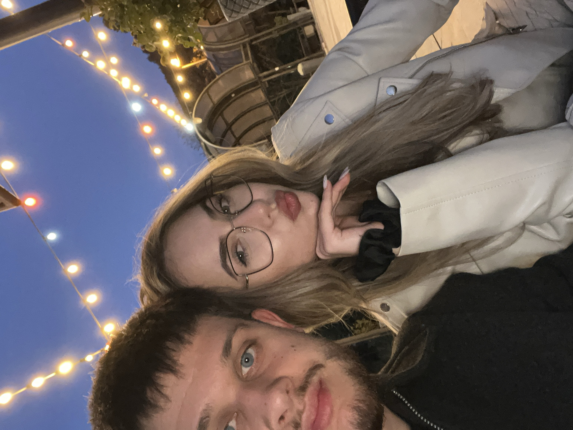 **Description:**
The image captures a selfie of a man and a woman, seemingly at dusk or twilight. The man is in the foreground, his face partially visible, showcasing a light beard and blue eyes. The woman is positioned slightly behind and above him, wearing glasses and a light-colored jacket. She makes a kiss gesture towards the camera.
In the background, a string of warm-toned lights is visible against the darkening sky. Behind the couple, there's an architectural structure, possibly a part of a building or an outdoor venue. Based on the architectural features visible in the background, the image might be taken in the vicinity of "Tsentralen ploshtad", a main square within Blagoevgrad that might include a building or an outdoor venue with these characteristics.