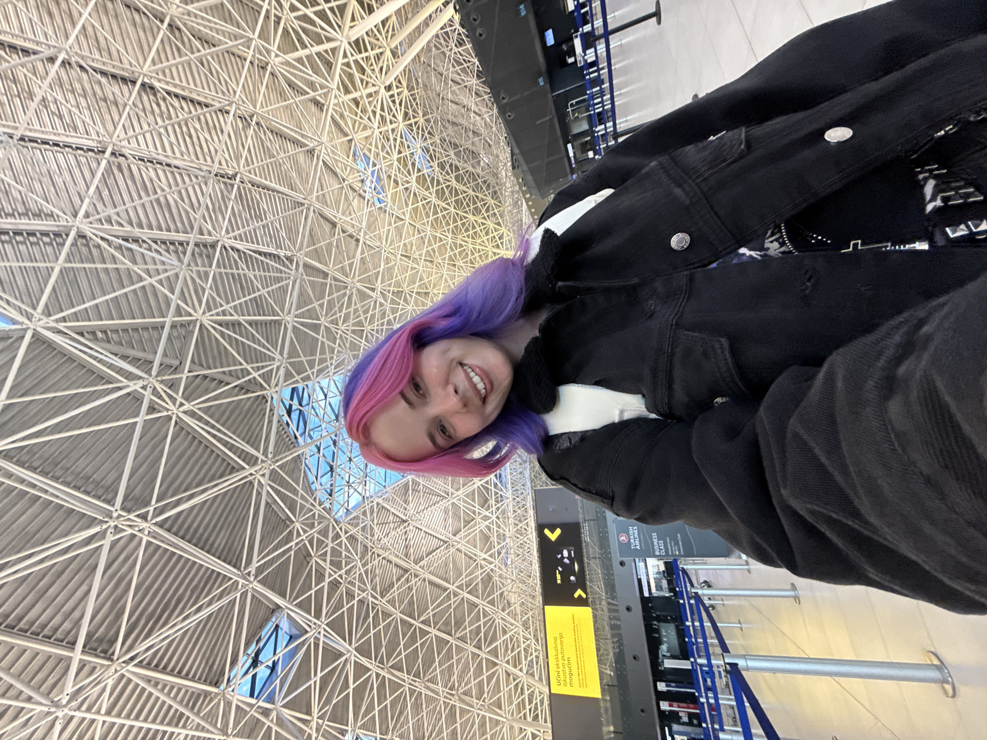 A smiling young woman with bright pink and purple hair stands at Zagreb Airport (ZAG) in the city of Velika Gorica, Croatia. She is wearing a black denim jacket and the airport's metal grid ceiling is visible above her. Airport signage with black lettering on a yellow background is in the background. Stanchions with blue straps are also in the background.