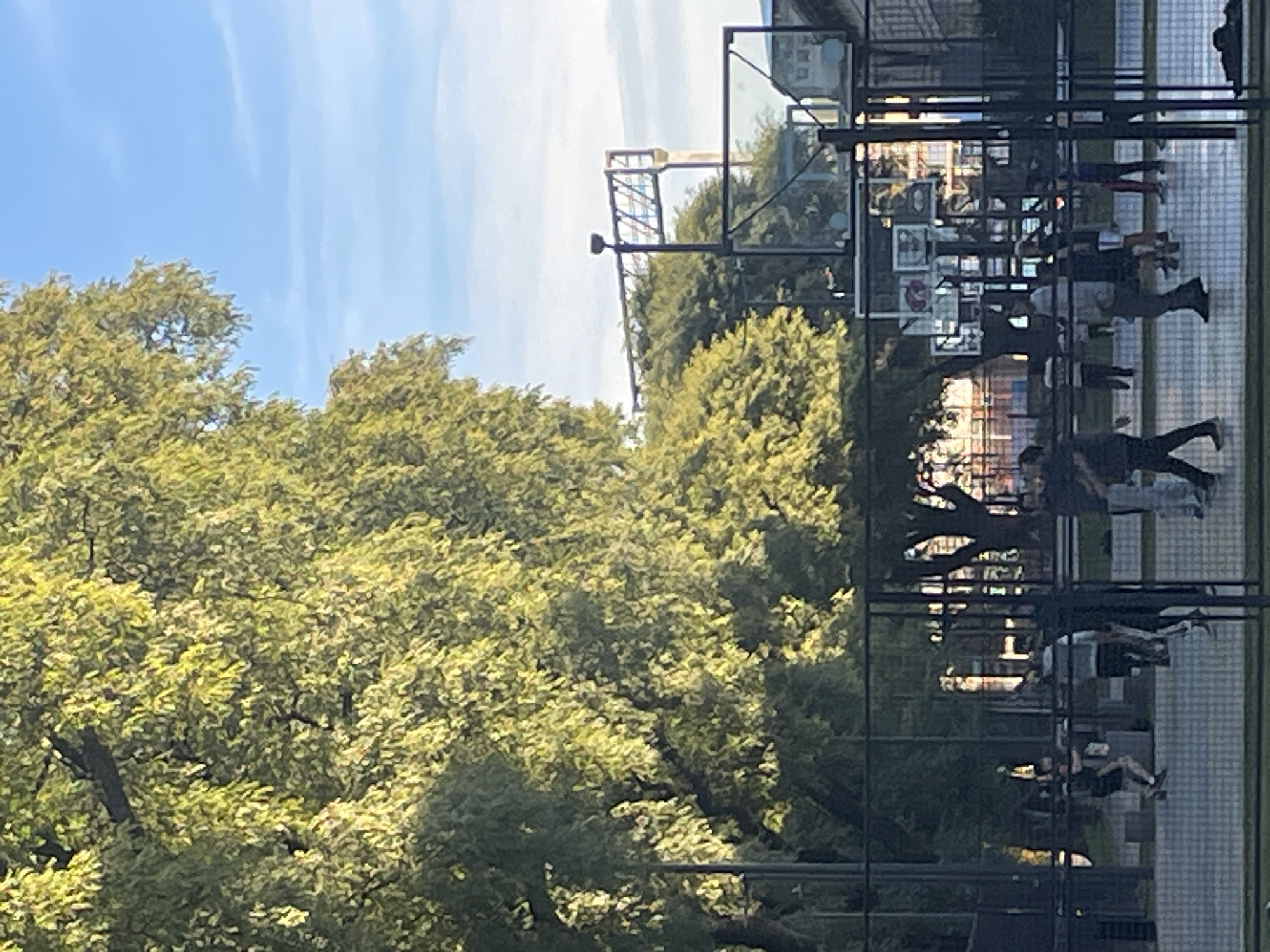The image depicts a group of people walking within a fenced-in area in Buenos Aires, Argentina. The background includes green trees and a sky with light clouds. A tall structure with lights is visible above the trees. The fenced-in area is paved and possibly serves as a public space. The people are walking and interacting in an everyday manner.