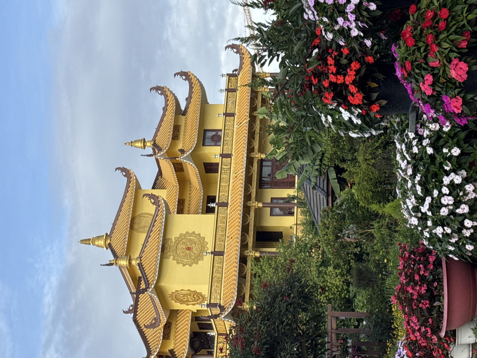 The image shows a tall, yellow Buddhist temple with ornate golden roofs and decorations, standing amidst lush green foliage and vibrant flower planters. The temple appears to have multiple levels, adorned with traditional architectural details. To the right of the temple, there are large flower planters with a variety of colorful flowers, including red, pink, and white varieties. The sky is overcast with blue patches. The temple is located in Da Lat, Vietnam.