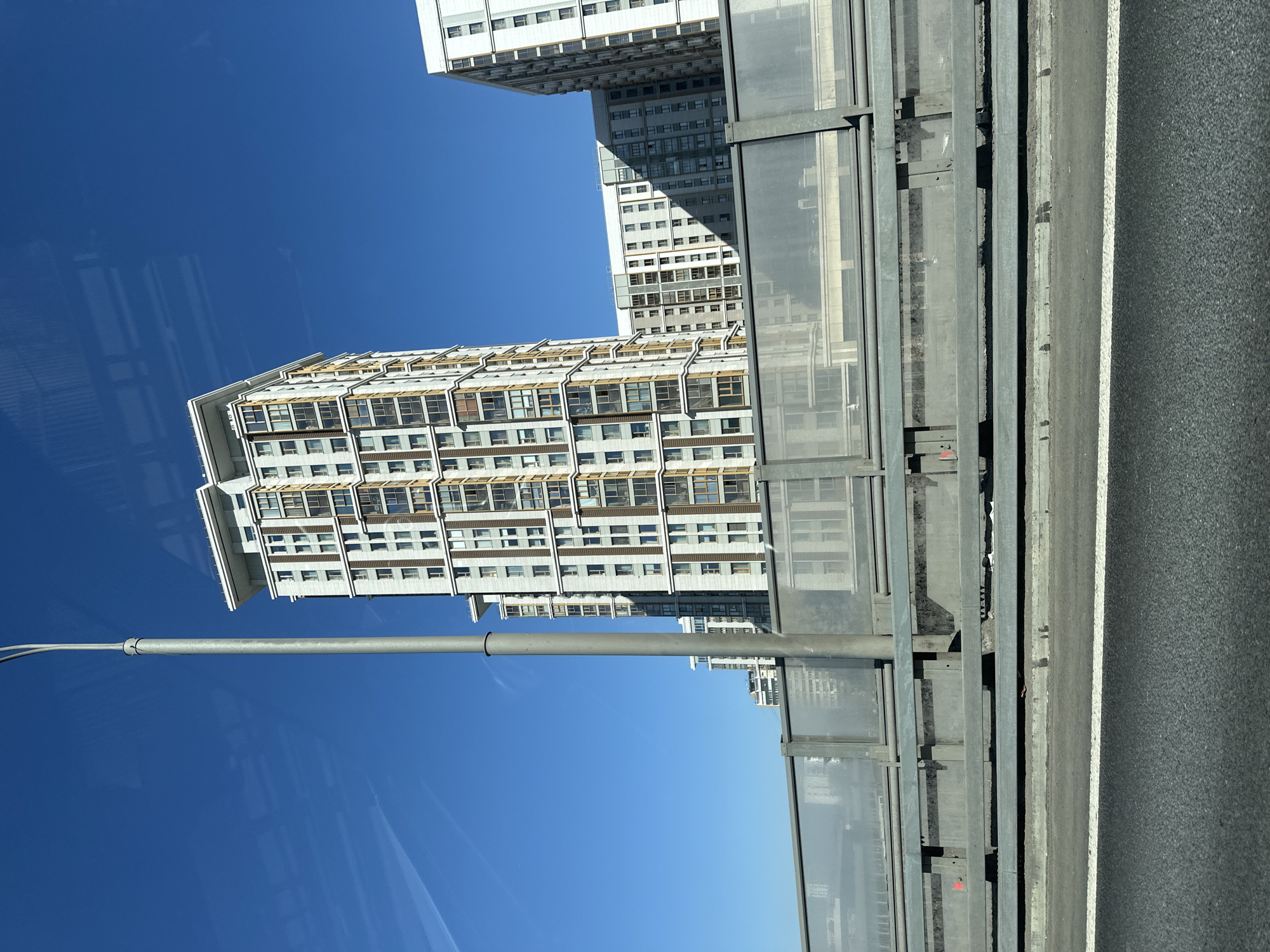 The image shows a view from a bridge in Moscow, Russia. A modern, high-rise residential building with numerous windows is visible against a clear blue sky. A sound barrier runs along the bridge edge, and a lamppost is partially visible on the left side of the frame. The road surface is asphalt with painted lane markings.