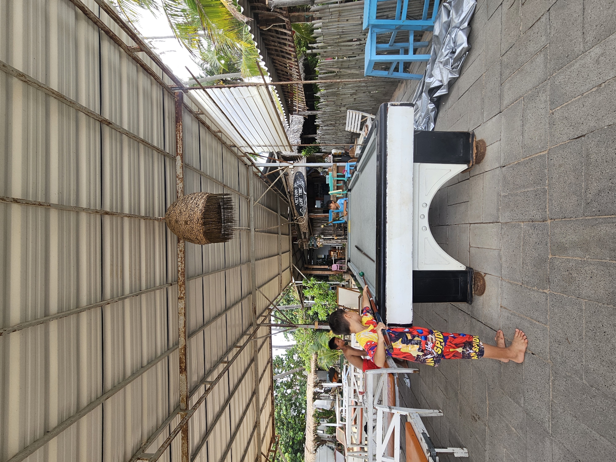 The image depicts an outdoor billiards table setup at a location in Mũi Né, Vietnam. A young boy wearing colorful shorts patterned with Spider-Man is actively engaged in playing billiards, positioned on the left side of the table and striking a ball with a cue. To his left, another boy in red shorts stands near the table. The billiards table is white with black accents and rests on the ground. The entire area is covered by a corrugated metal roof supported by metal beams, suggesting it is part of an open-air establishment or resort. In the background, there are thatched-roof structures and lush green palm trees, characteristic of a tropical environment. The ground is paved with grey rectangular tiles. Blue stools are visible to the right of the billiards table, alongside a bamboo fence. The scene appears to be a recreational area at a beachside resort or restaurant in Mũi Né.