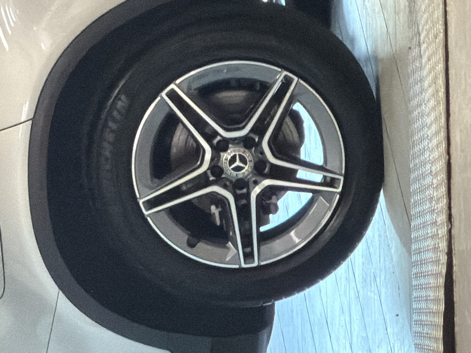 The image displays a close-up of the passenger-side wheel of a white or light-colored Mercedes-Benz vehicle, located in Aalsmeer, Netherlands. The wheel consists of a five-spoke, two-tone alloy rim with a Mercedes-Benz star logo centered on the hubcap. The tire is a Michelin brand and appears significantly underinflated or flat, exhibiting a distinct flattening where it contacts the ground surface. A dark, textured fender arch trim is visible above the tire. Through the spokes of the wheel, a brake rotor and caliper assembly are discernible. The vehicle is positioned on a light blue or pale grey floor with visible parallel lines, suggesting a tiled or planked surface. In the bottom foreground, a section of a light-colored, woven mat or carpet runner is present. No individuals or active events are depicted.