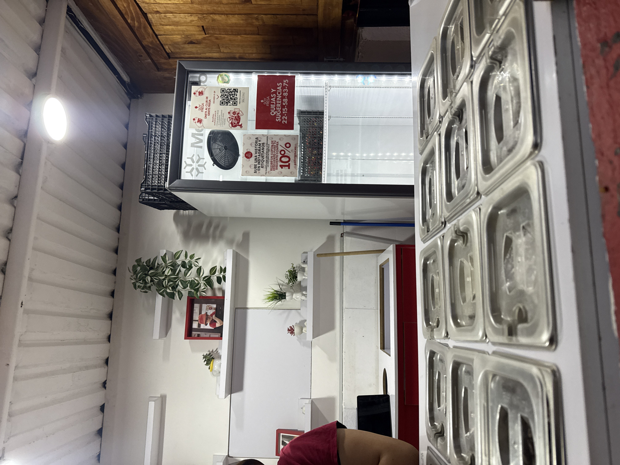 Here is a description of the image:
The image shows the interior of a commercial establishment, possibly a restaurant or food service business, located in Puebla, Mexico.
The location is suggested by the signage, which features the name "FRESA" and local contact information (22-15-58-83-75) that is specific to Puebla. The establishment's walls are white. White shelves hold decorations, including plants and framed pictures.
A commercial refrigerator is visible, containing promotional materials, including advertisements and contact information. To the right are stainless steel trays, suggesting food preparation or storage areas. Part of a person is visible in the lower center of the image. The person's skin is visible, and they are wearing a burgundy shirt. The image is rotated, as the floor-to-ceiling view is not horizontal.