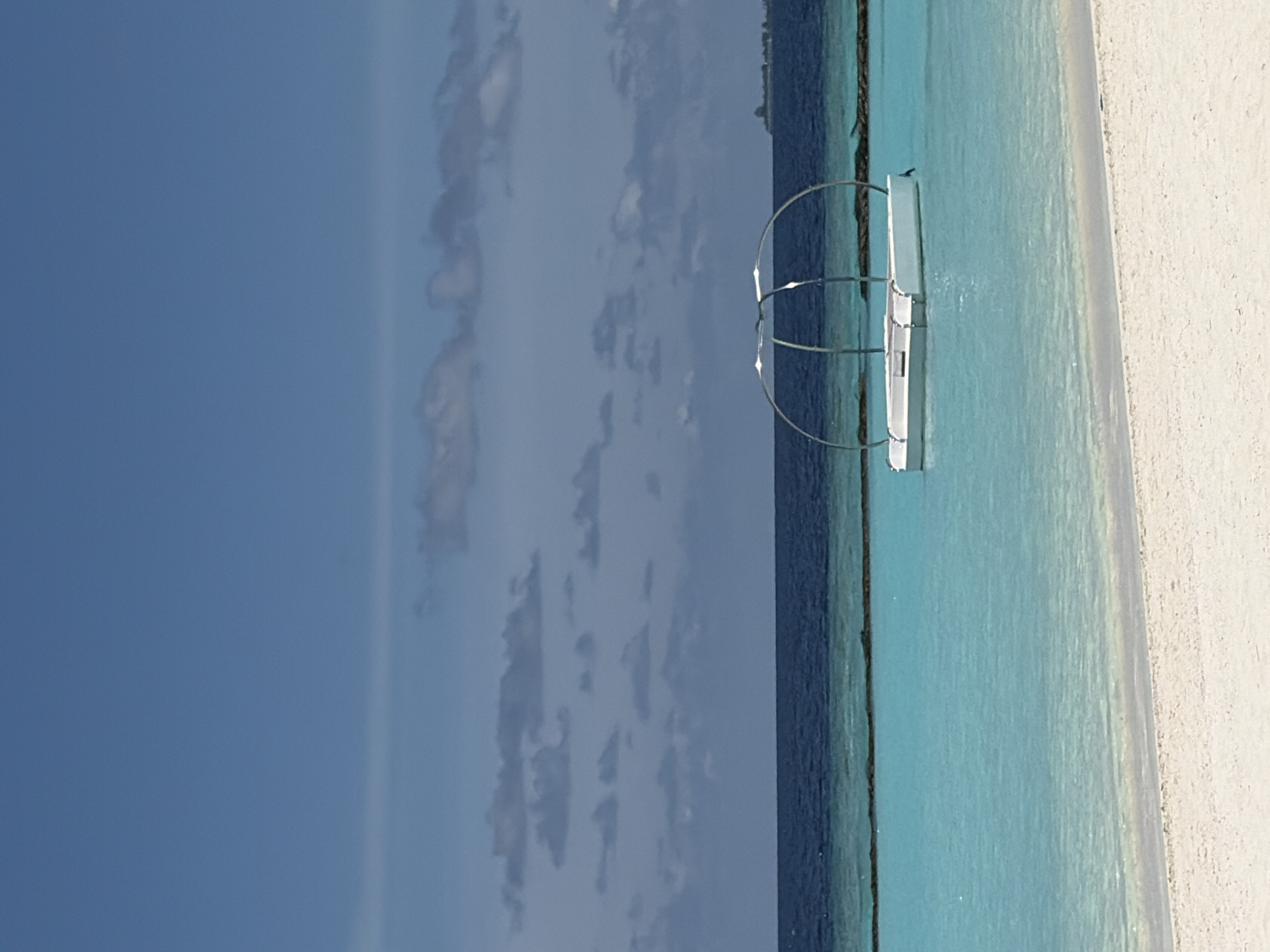 The image depicts a serene beach scene in Halaveli, Maldives. In the foreground, a pristine white sand beach meets the tranquil, turquoise waters of the ocean. The water transitions to a deep blue further out, meeting the horizon. A small, white floating structure with a curved metal frame sits on the water, appearing to be a decorative element or a small pontoon. In the distance, a faint outline of land can be seen along the horizon, likely another island. The sky is a clear blue, dotted with a few wispy clouds, suggesting fair weather. The overall atmosphere is peaceful and idyllic, characteristic of a tropical resort.