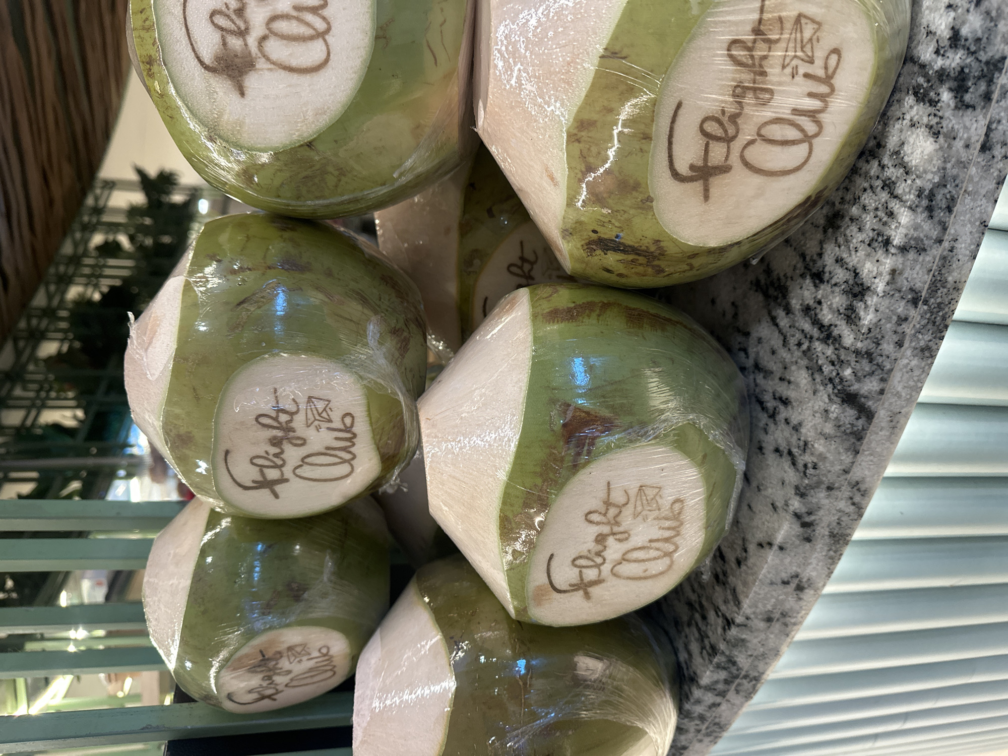 A stack of young coconuts, wrapped in clear plastic, are displayed on a speckled grey countertop with a light blue ribbed base. Each coconut has a white, circular label with brown text that appears to say "Flight Club" and a small drawing of a paper airplane. The coconuts are partially opened, revealing their white flesh. The background suggests an indoor setting with green metal shelving and tropical foliage visible. The overall scene is clean and suggests a commercial display, likely a beverage stand or market stall in Kuta, Indonesia, given the context of the original prompt.
