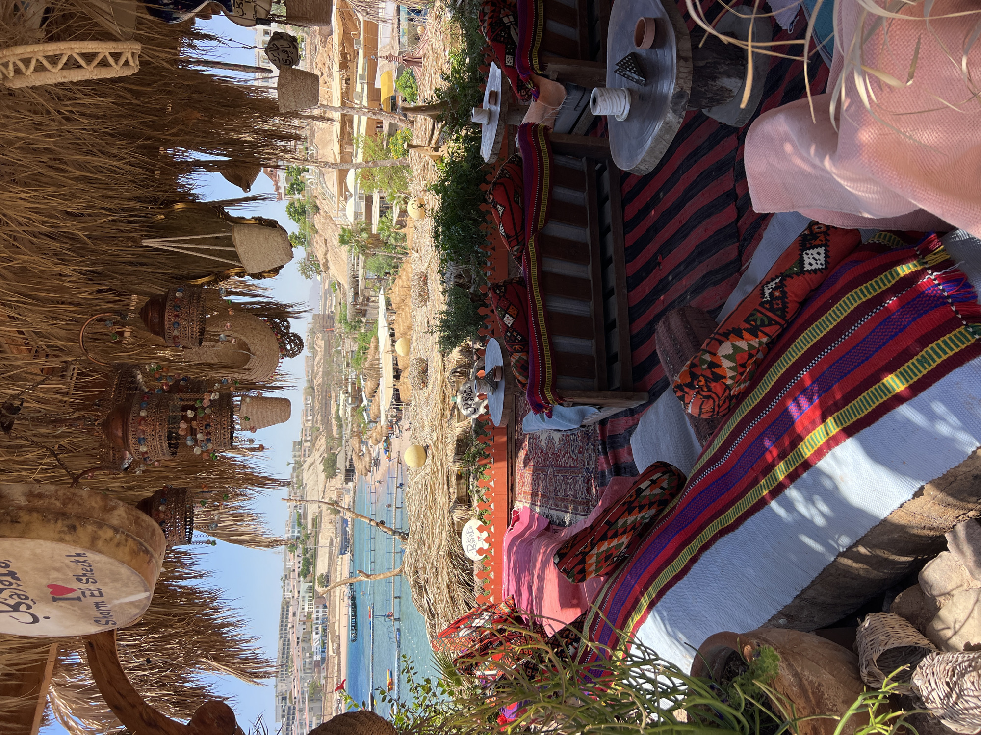 This is an outdoor seating area at a beach resort in Sharm El Sheikh, Egypt. The area is covered by a thatched roof and decorated with traditional Middle Eastern textiles and lanterns. In the foreground, there are several comfortable seating arrangements with plush cushions and colorful rugs. In the background, a beach with sunbathers and umbrellas can be seen, along with the blue waters of the Red Sea and distant hills dotted with buildings. The overall atmosphere is relaxed and inviting, typical of a vacation destination.