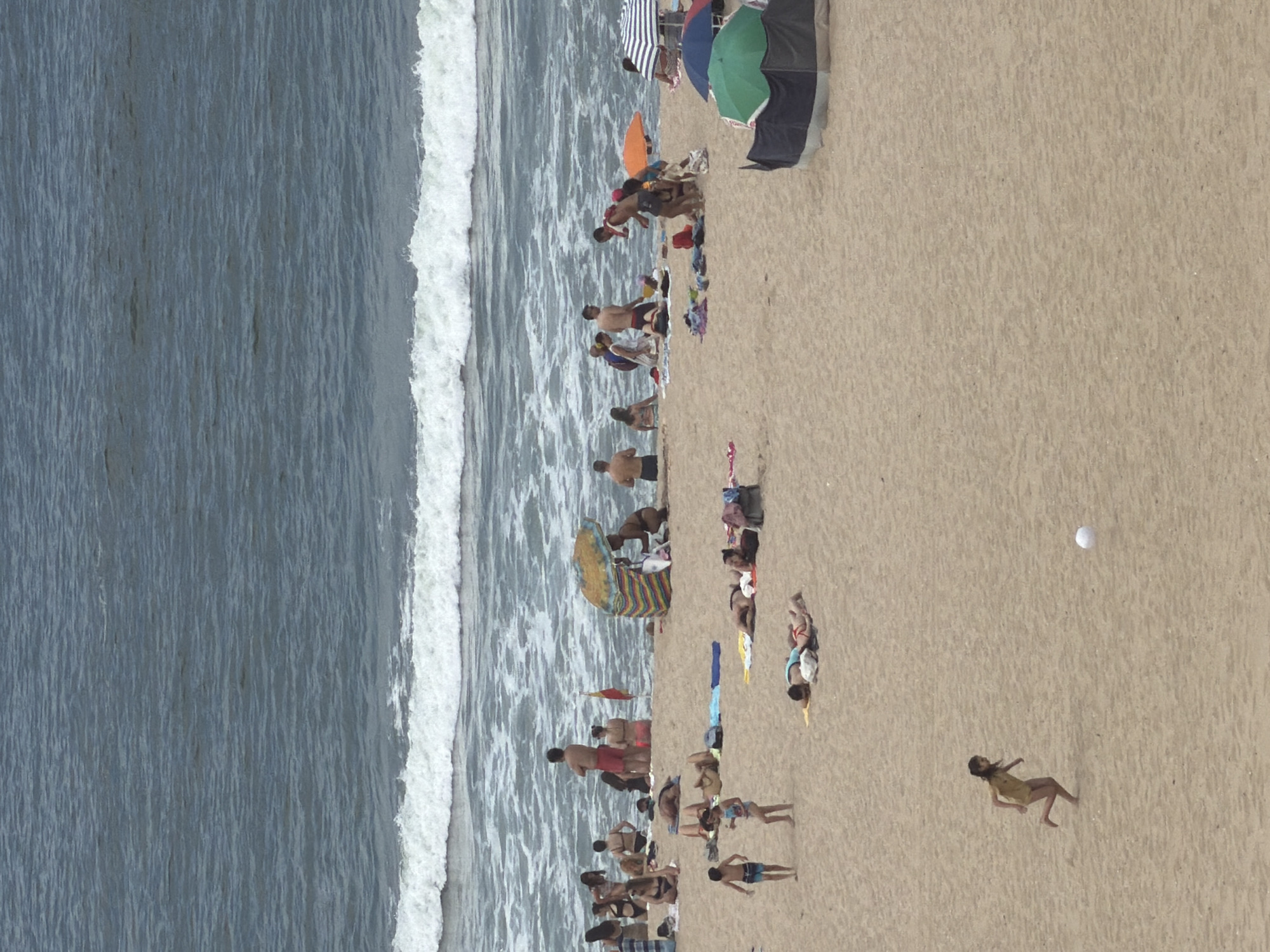 The image depicts a crowded beach scene in Espinho, Portugal, with the Atlantic Ocean in the background. The water is choppy with white waves breaking near the shore. On the sandy beach, numerous people are engaged in various activities. Some are sunbathing on towels and blankets, while others are sitting or standing. Several beach umbrellas and a small beach tent provide shade. A young girl in a yellow swimsuit is running across the sand, kicking a white ball. Other individuals are walking or gathered in groups. The overall atmosphere suggests a busy summer day at the beach.