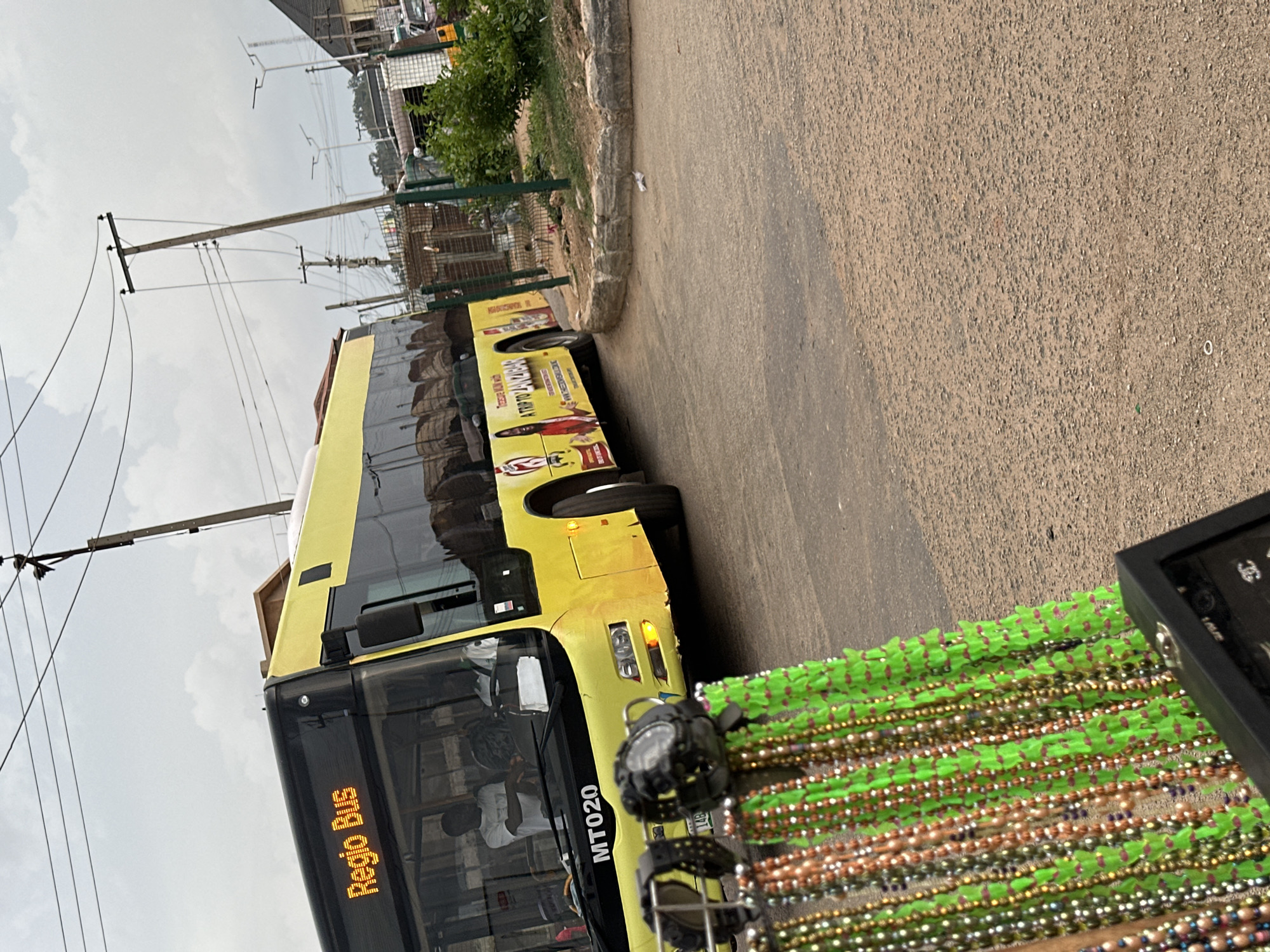 A yellow and black Regio Bus with the designation MT020 is parked on a dusty road in Bukuru, Nigeria. The bus has advertisements on its side, one of which depicts a person with the text "A TRIP TO ZANZIBAR." In the foreground, a display rack holds various necklaces and watches for sale. The road surface is a mix of asphalt and gravel, with scattered debris. Power lines are visible overhead. The sky is overcast.
