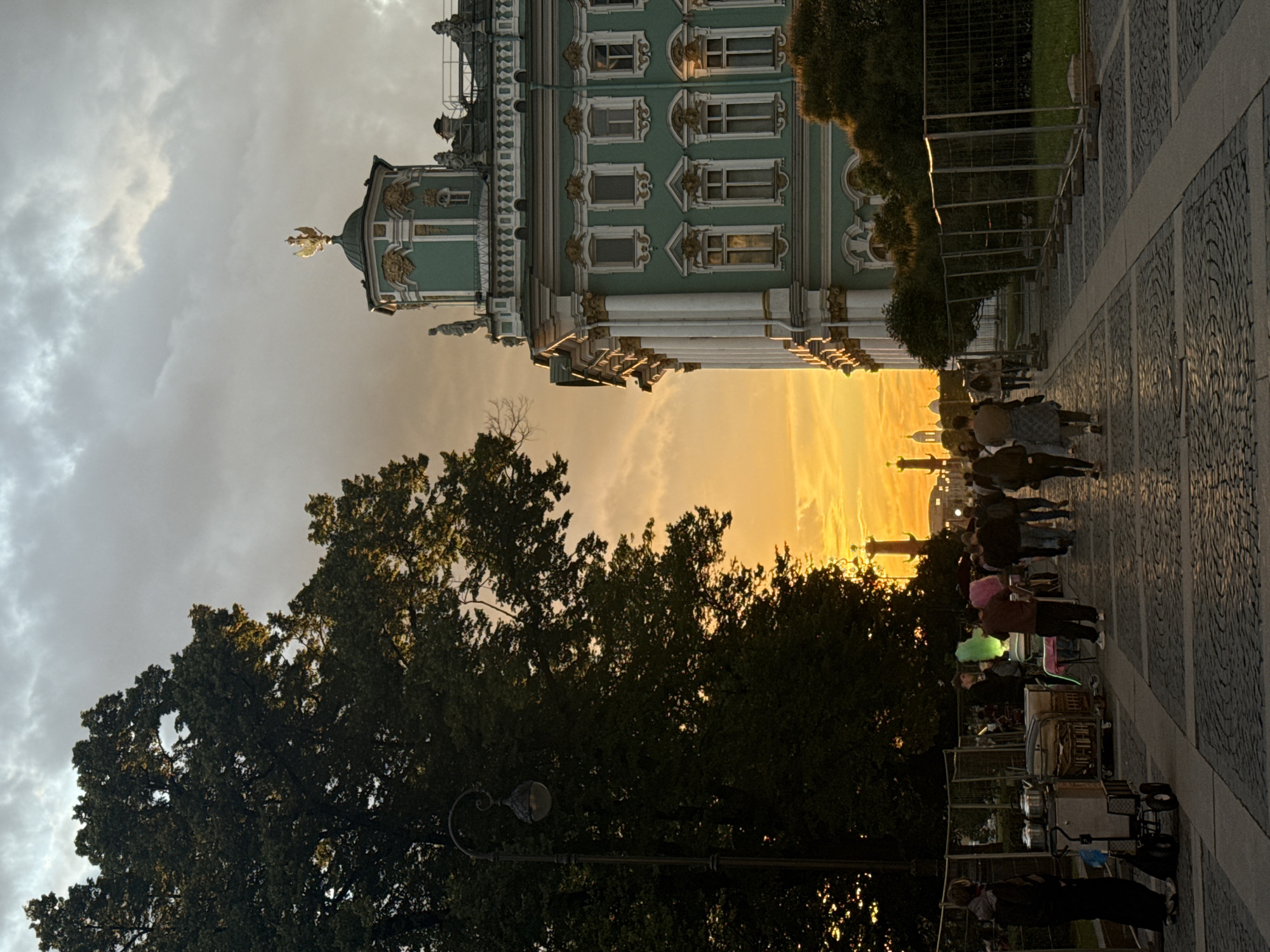 This photograph captures a dramatic sunset over a grand architectural setting, likely in Saint Petersburg, Russia, given the style of the building and the presence of what appears to be a column in the distance. The sky is filled with vibrant orange and yellow hues, with streaks of clouds illuminated by the setting sun, creating a warm and slightly hazy atmosphere.
In the foreground, a richly decorated building with green facades, white columns, and ornate gold detailing stands prominently. The architecture suggests a historical palace or museum. To the right, a cobblestone walkway is lined with trees and a black metal fence. Several people are visible on the walkway, some walking, while others appear to be gathered or engaged in activities, possibly street vendors with carts and colorful items. One individual is wearing a bright pink wig.
The overall mood is serene and picturesque, amplified by the beautiful natural light. The time of day is clearly evening, marked by the intense sunset. The weather appears to be clear, allowing for the spectacular display of colors in the sky. The image evokes a sense of cultural tourism and the beauty of an urban landscape during golden hour. No visible text is present in the image.