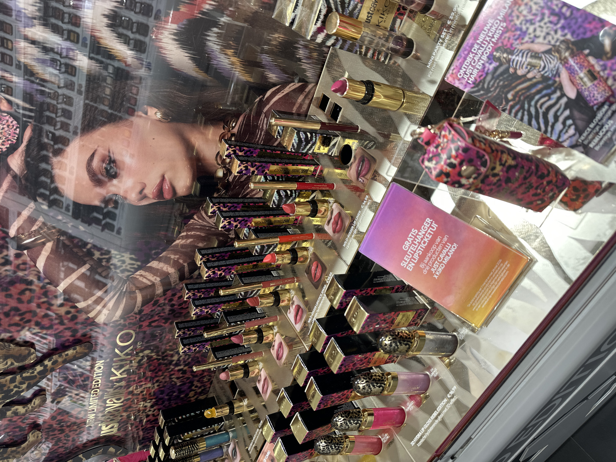 The image depicts a Kiko Milano makeup display, featuring the "Just Cavalli x Kiko Milano" limited edition collection. The display includes lipsticks, lip liners, lip glosses, and other beauty products with leopard print packaging. There is also a promotional sign offering a free keychain and lipstick holder with the purchase of three Just Cavalli x Kiko Milano products. Another sign advertises new Just Cavalli x Kiko Milano hair and body mists. The display features a model in a leopard-print dress, presumably advertising the makeup line. It can be inferred that this display is located in a store, possibly in Amsterdam, Netherlands, given the inclusion of Dutch language.