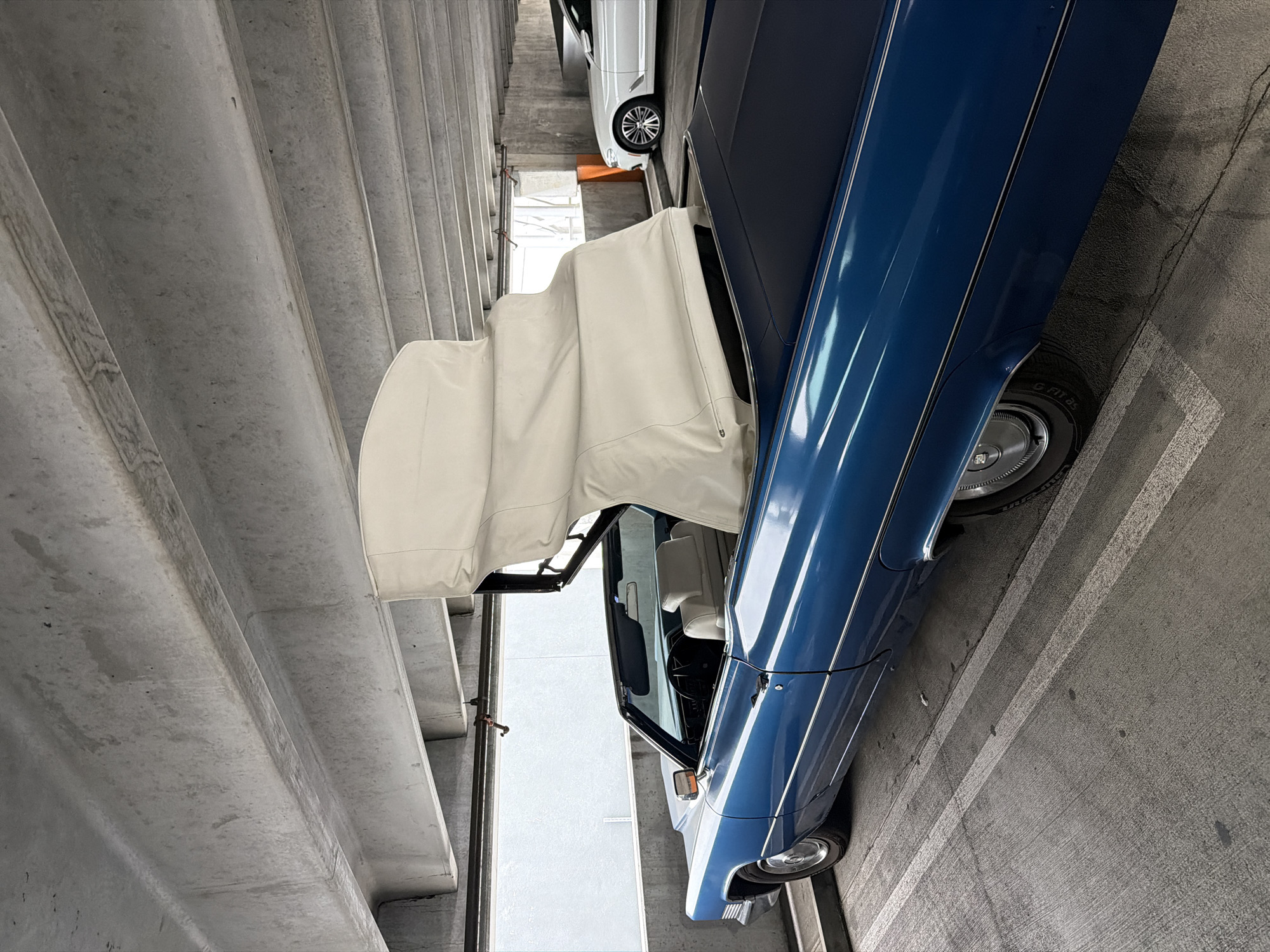 An angled, eye-level, medium outdoor shot shows the rear right quarter panel of a vintage blue Cadillac convertible with its white convertible top partially raised in a concrete parking garage. The car is parked in a marked parking space with white lines on the concrete floor. The car's interior is visible, featuring white leather seats. Behind the Cadillac, a white modern sedan is parked in an adjacent space. The ceiling of the parking garage is visible above, constructed of concrete beams and slabs. Natural light filters in from an opening at the far end of the garage, illuminating the scene. The overall environment suggests a public or private parking facility in a city.