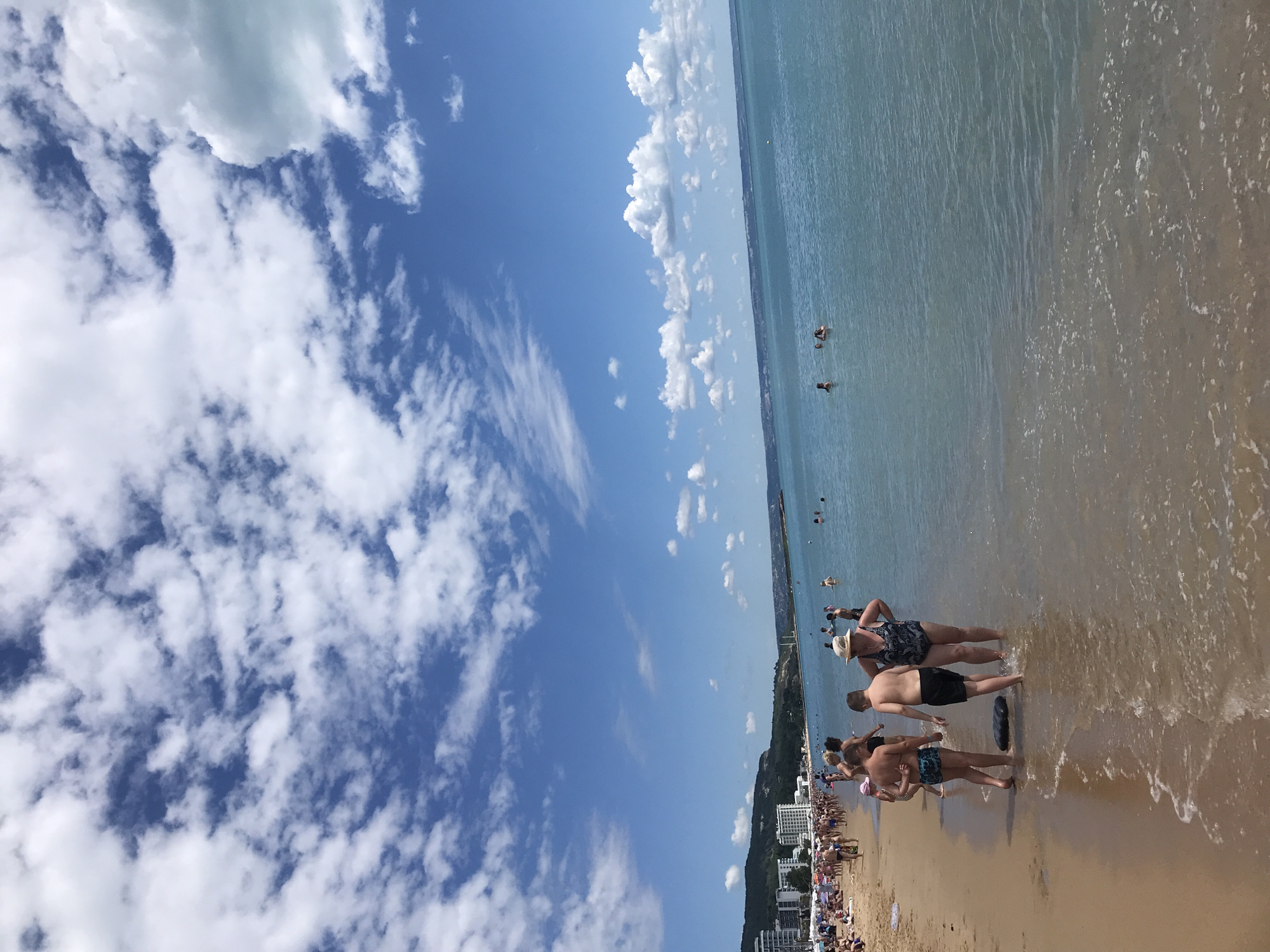 This image depicts a crowded beach scene in Varna, Bulgaria, likely on a sunny day given the blue sky dotted with white clouds. The foreground shows a sandy beach with several people walking at the edge of the water. Among them are two young boys in swimwear and a woman wearing a patterned swimsuit and a hat. The water is clear and a gentle wave is washing onto the shore. In the background, a long stretch of beach is visible, lined with sunbathers and a promenade with buildings and hotels behind them. Further in the distance, a coastline with hills covered in vegetation can be seen, along with a bridge or pier extending into the sea. Several people are swimming or wading in the turquoise water. The overall impression is a typical summer day at a popular seaside resort.