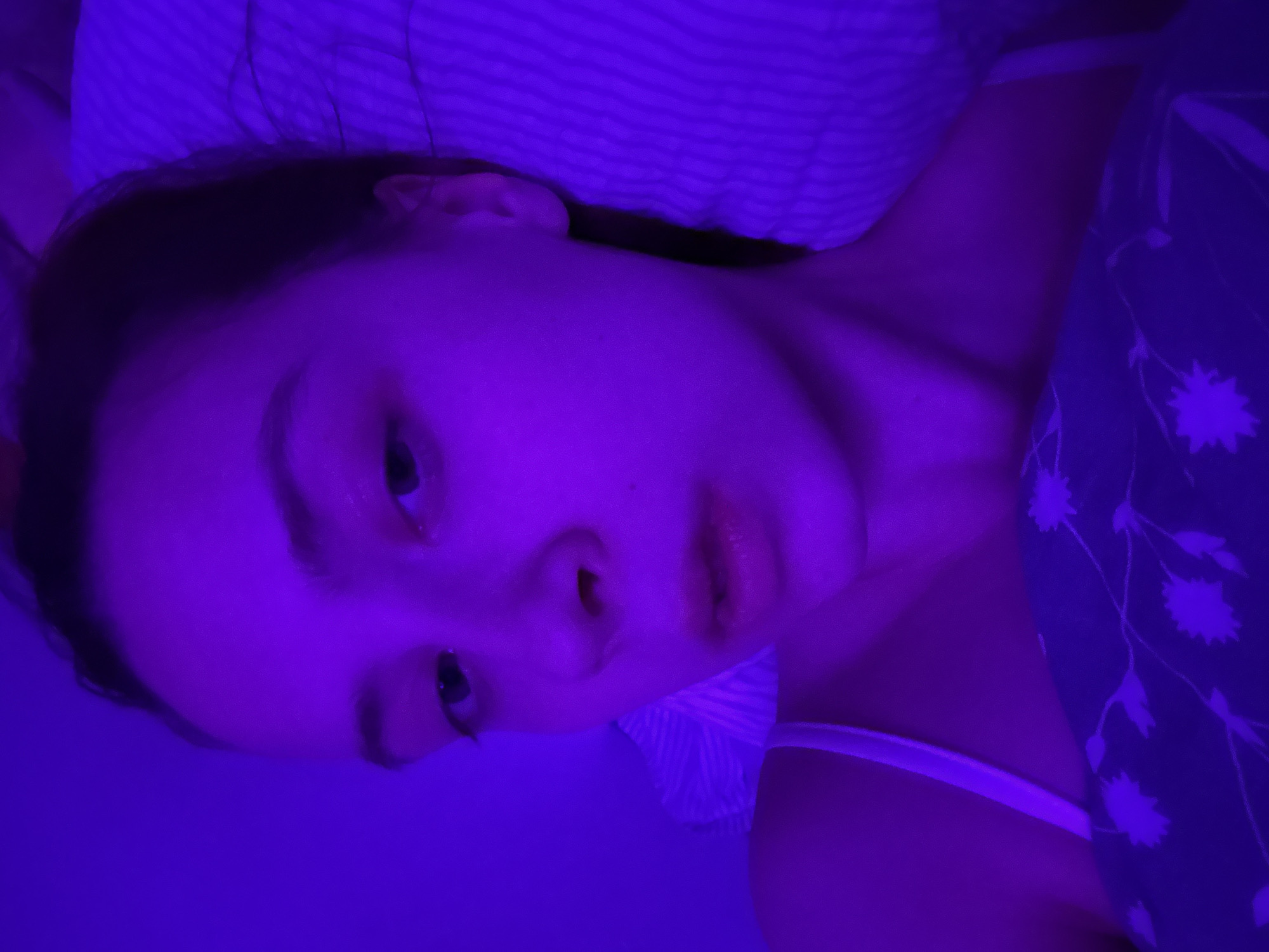 A young woman lies in bed under a blue light. She appears to be lying on her side, her head on a striped pillow. Her face is visible, with her eyes open. She has dark hair pulled back from her face. She is wearing a white tank top under a dark blue shirt with a floral pattern.
The location is unknown; Прикамский, Russia is not visible or inferable from the image.