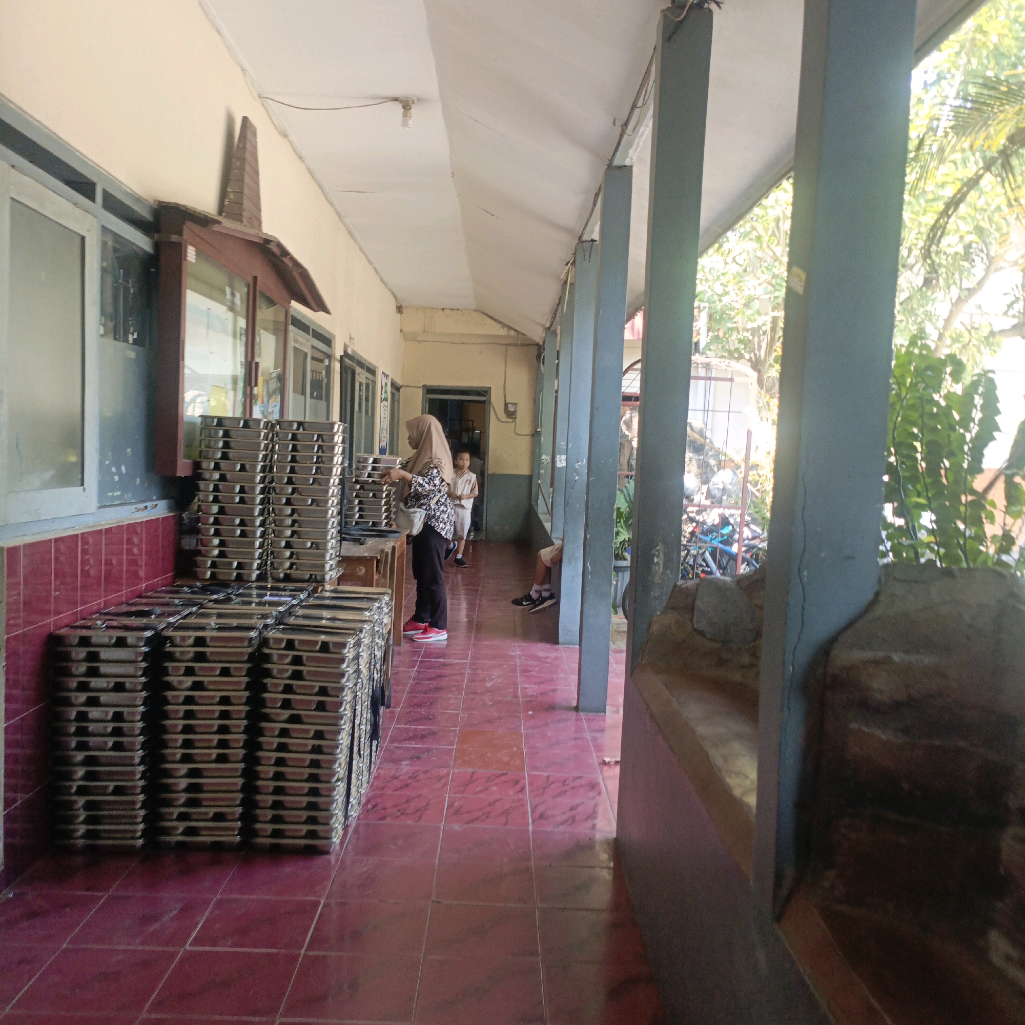 The image depicts a hallway at a school in Salatiga, Indonesia. Piles of stacked metal trays are visible on the left side of the hallway. A woman wearing a hijab and a patterned shirt is standing near the trays, and a young person in a school uniform is walking further down the hallway. The hallway has a tiled floor and is lined with pillars on the right side. Sunlight streams into the hallway from the right, illuminating some of the area and the greenery outside. A bicycle can be seen parked in the background.