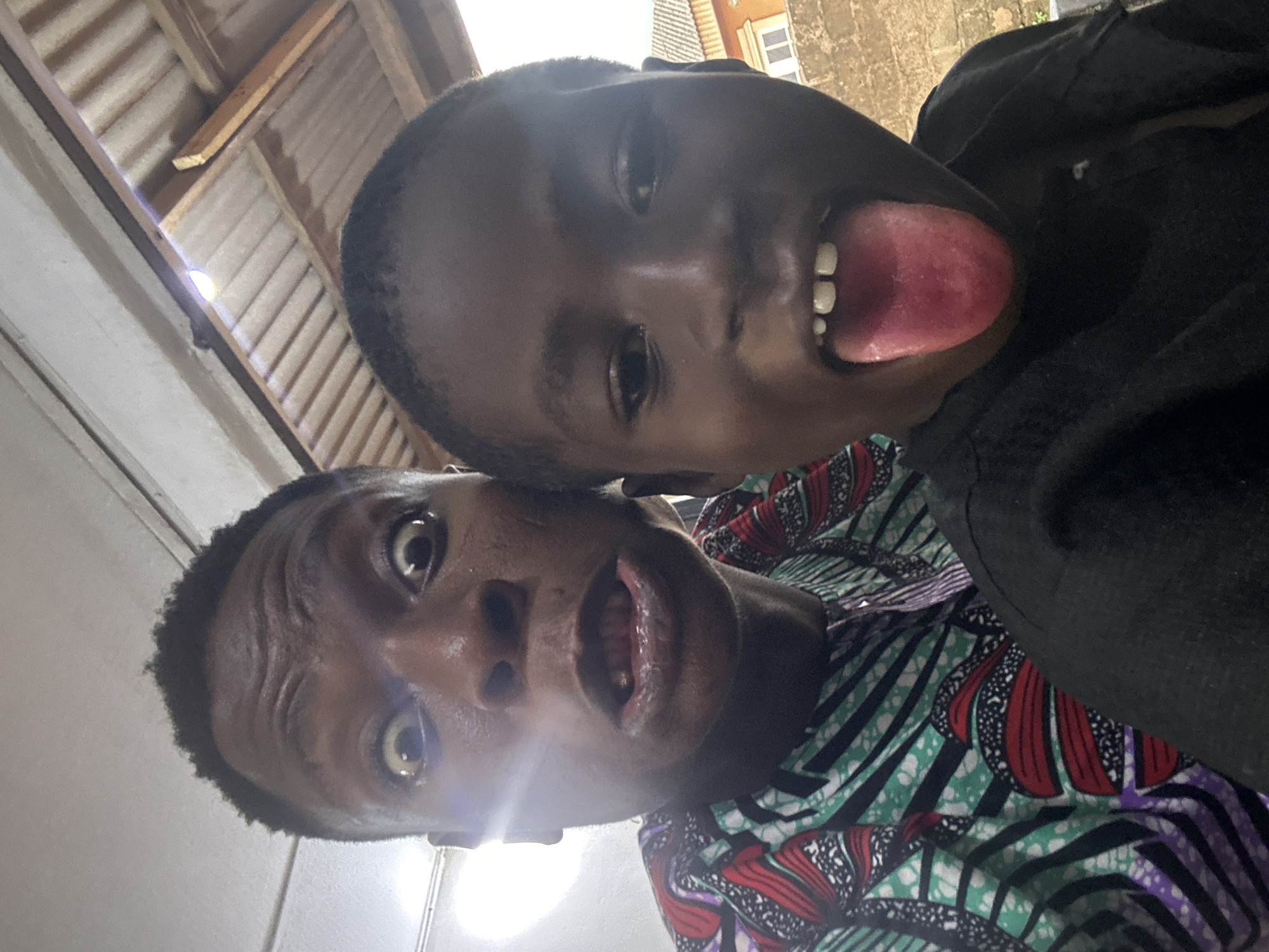 Two dark-skinned individuals are captured in a close-up, tilted selfie. The person in the background is sticking their tongue out. The person in the foreground has wide eyes and an open mouth. One is wearing a patterned garment and the other is wearing a black shirt. The setting appears to be indoors, with a ceiling visible in the background. The city and state are unknown.