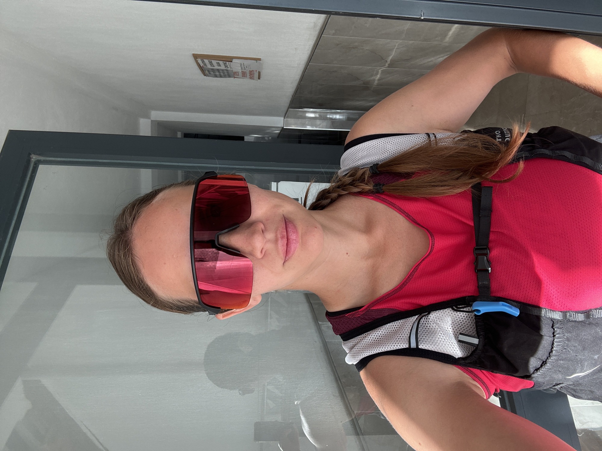A woman in activewear and mirrored sunglasses stands inside a building with a gray-framed glass door. She is wearing a red tank top under a black and gray hydration pack, her hair is styled in a braid. The background shows a tiled floor and a sign attached to the white ceiling. Given the context of the city, there are no distinct identifiers suggesting the exact location in Fethiye, Turkey.