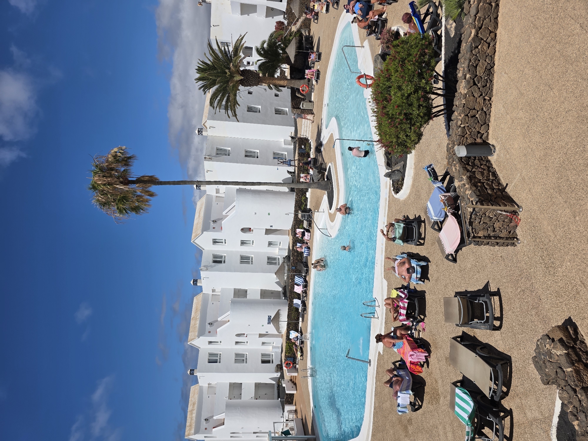 The image depicts a resort scene in Costa Teguise, Spain, likely on a sunny day. The primary focus is a bright blue swimming pool, curved and surrounded by a beige tiled deck. Several people are visible enjoying the water and lounging by the pool. Some are swimming or wading, while others are relaxing on lounge chairs, some with towels and books. A few palm trees and green bushes add to the tropical atmosphere. White, multi-story resort buildings with visible balconies and windows line one side of the pool area.
The setting appears to be a holiday resort, given the presence of the pool, lounge chairs, and architecture. The time of day is likely midday, as indicated by the bright sunlight casting shadows. The weather is clearly sunny and warm, suitable for swimming and outdoor relaxation. The overall mood conveyed is one of leisure and vacation. The buildings have a distinct architectural style, with white walls and earth-toned roofs, which, along with the palm trees and the dry, rocky landscape visible in the background, are characteristic of the Canary Islands. No visible text is present in the image.