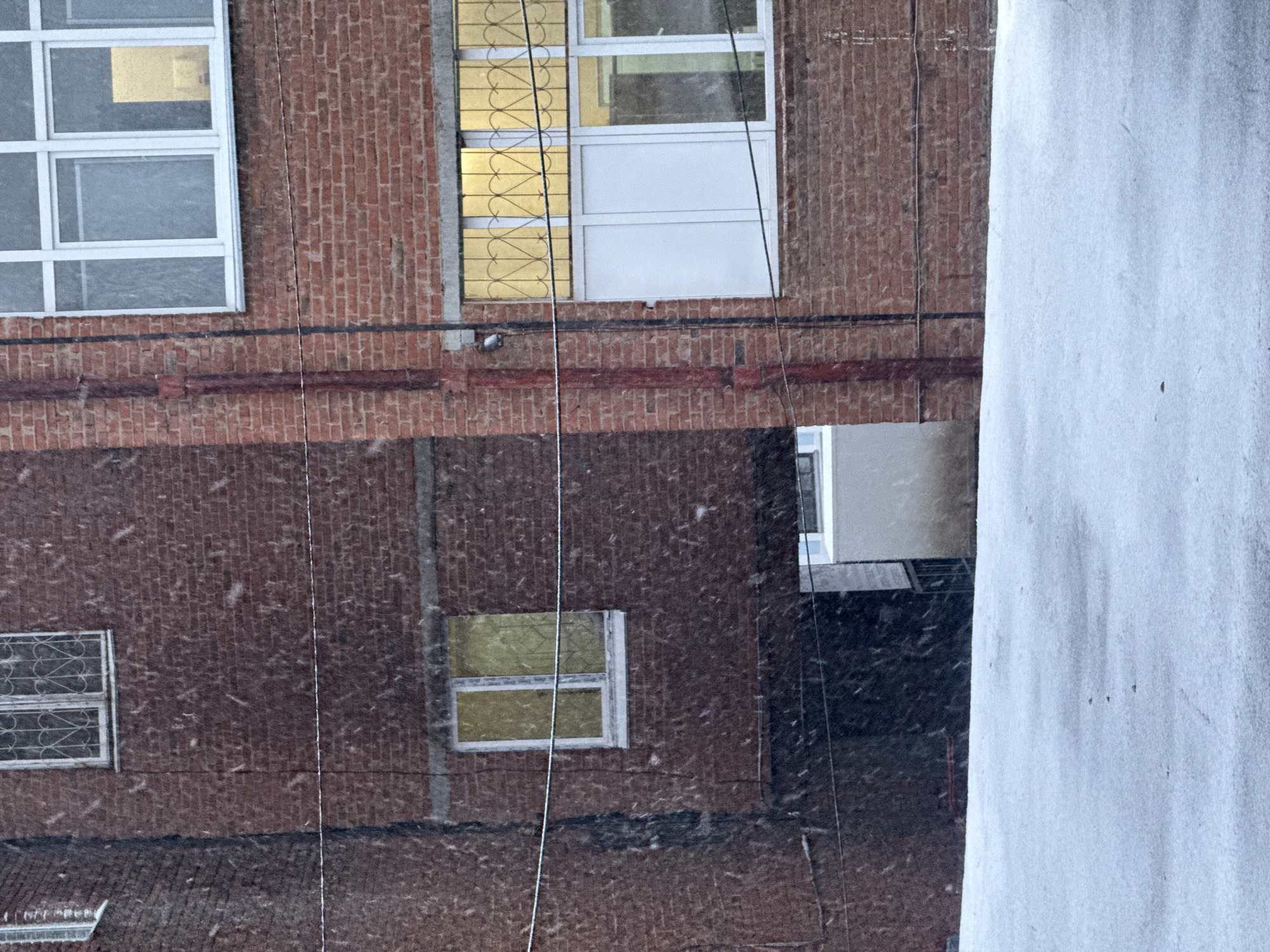 The image shows the exterior of a brick building with white-framed windows. It is snowing. In the foreground, there is a white wall partially obscuring the view. The architecture, including the brickwork and window styles, is typical of Soviet-era construction. The location is Votkinsk, Russia.