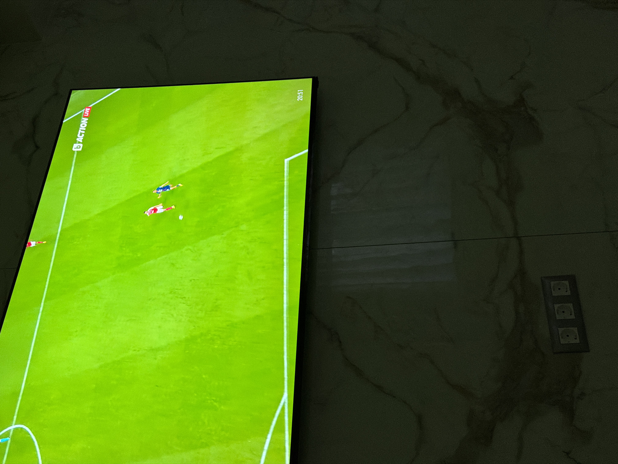 A television screen displays a live broadcast of a soccer game. The screen is mounted on a wall. A triple electrical outlet is visible on the wall to the right of the television. The time 20:51 is displayed at the top right of the screen. "ACTION LIVE" is displayed at the top left. The location is not identifiable from the image.